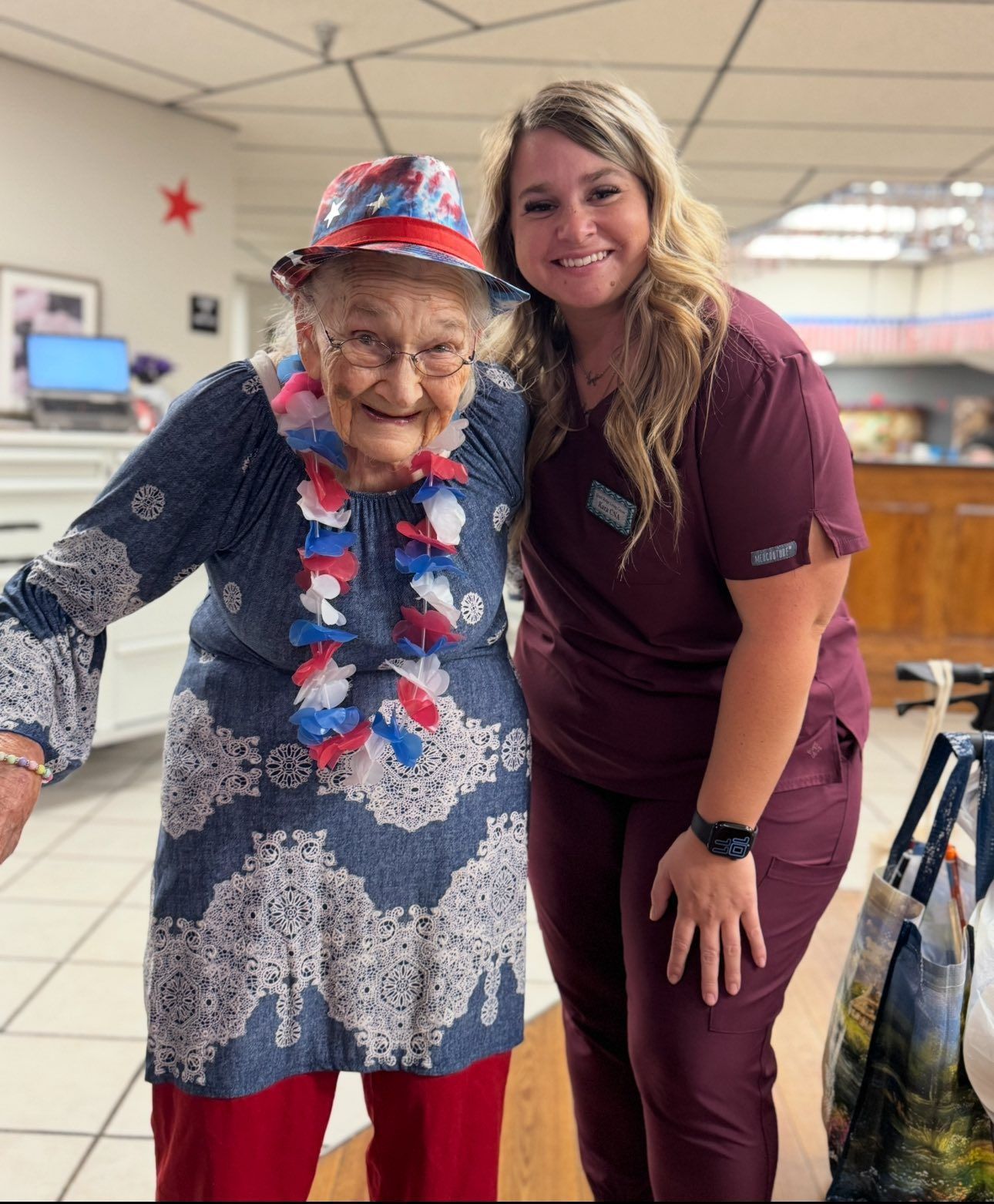 Elderly woman in festive attire with a caregiver smiling indoors, celebrating.