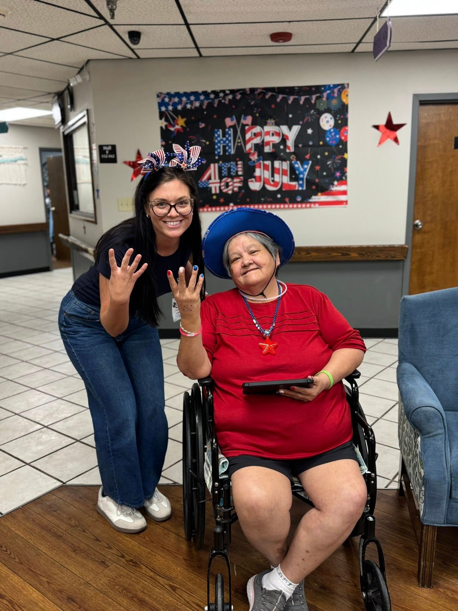 Woman in wheelchair and young woman celebrating July 4th. Both wear red, white, and blue, waving.