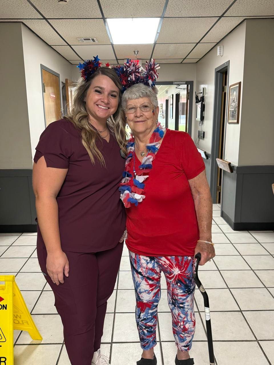 A woman in red and a staff member pose in a hallway, both wearing patriotic headwear.