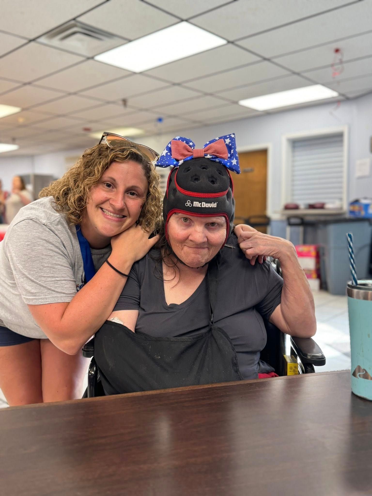 Woman smiles, poses next to another wearing a patriotic hat. Both are indoors, near a table.