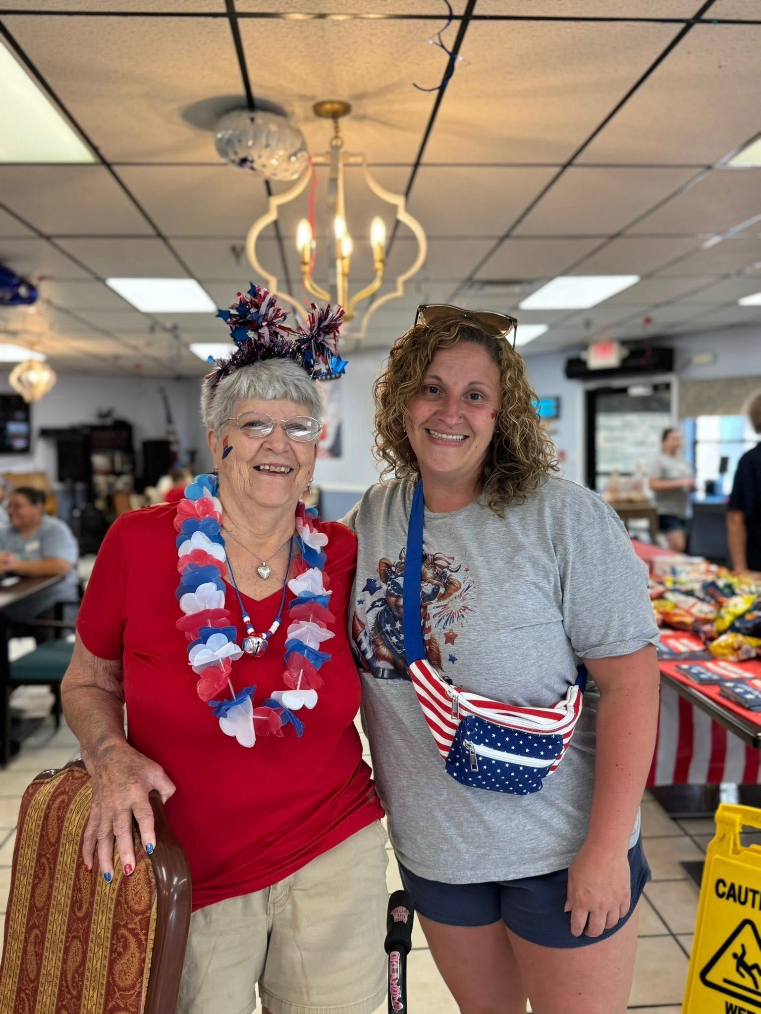 Two women pose together indoors, one in red, white, and blue attire; the other in gray shirt and shorts.