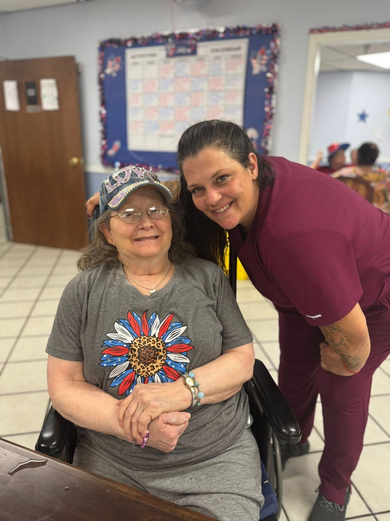 An elderly woman in a wheelchair smiles with a caregiver in a room.