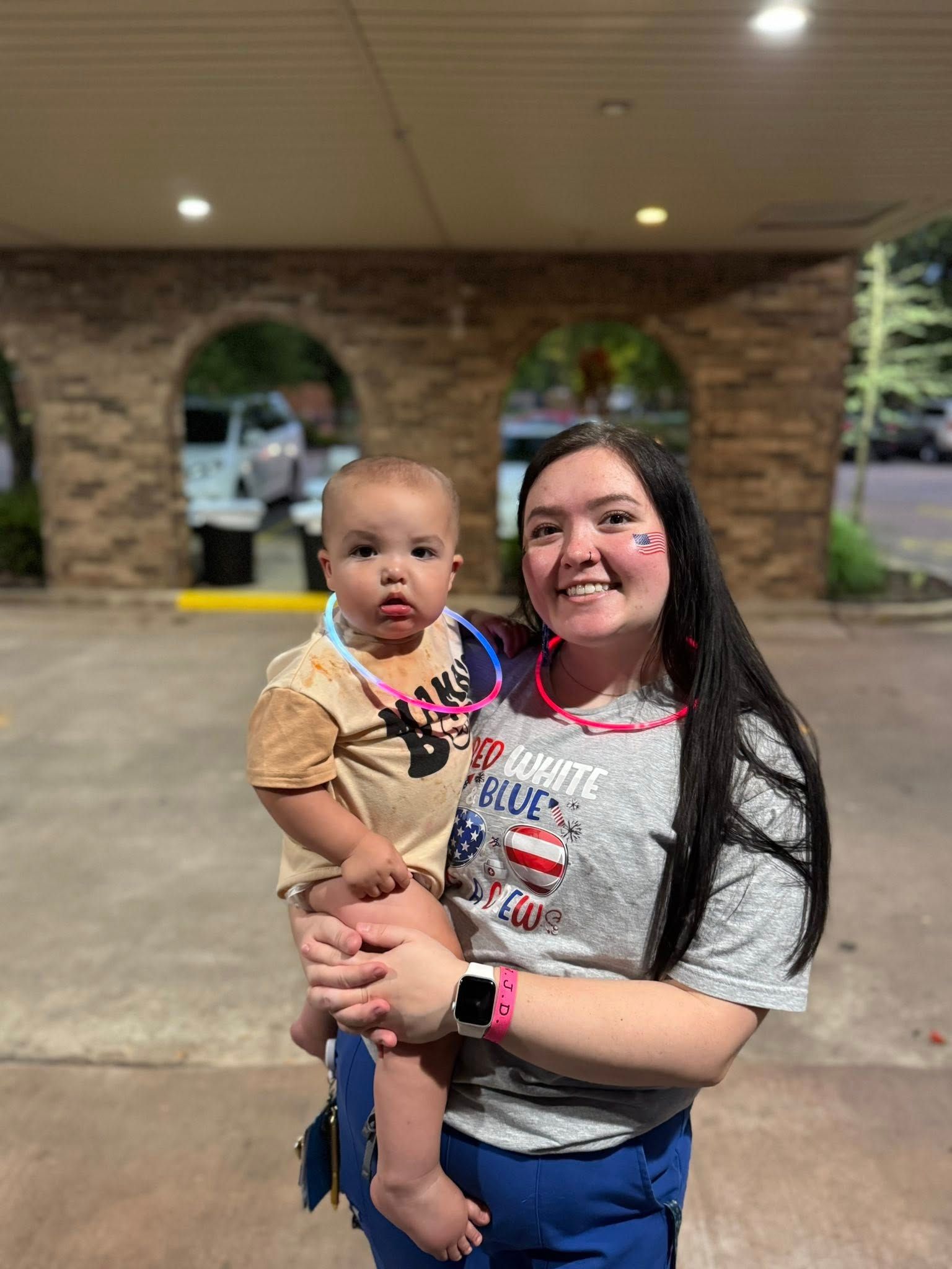 Woman holding baby, both wearing patriotic attire. Outside, near stone arches.
