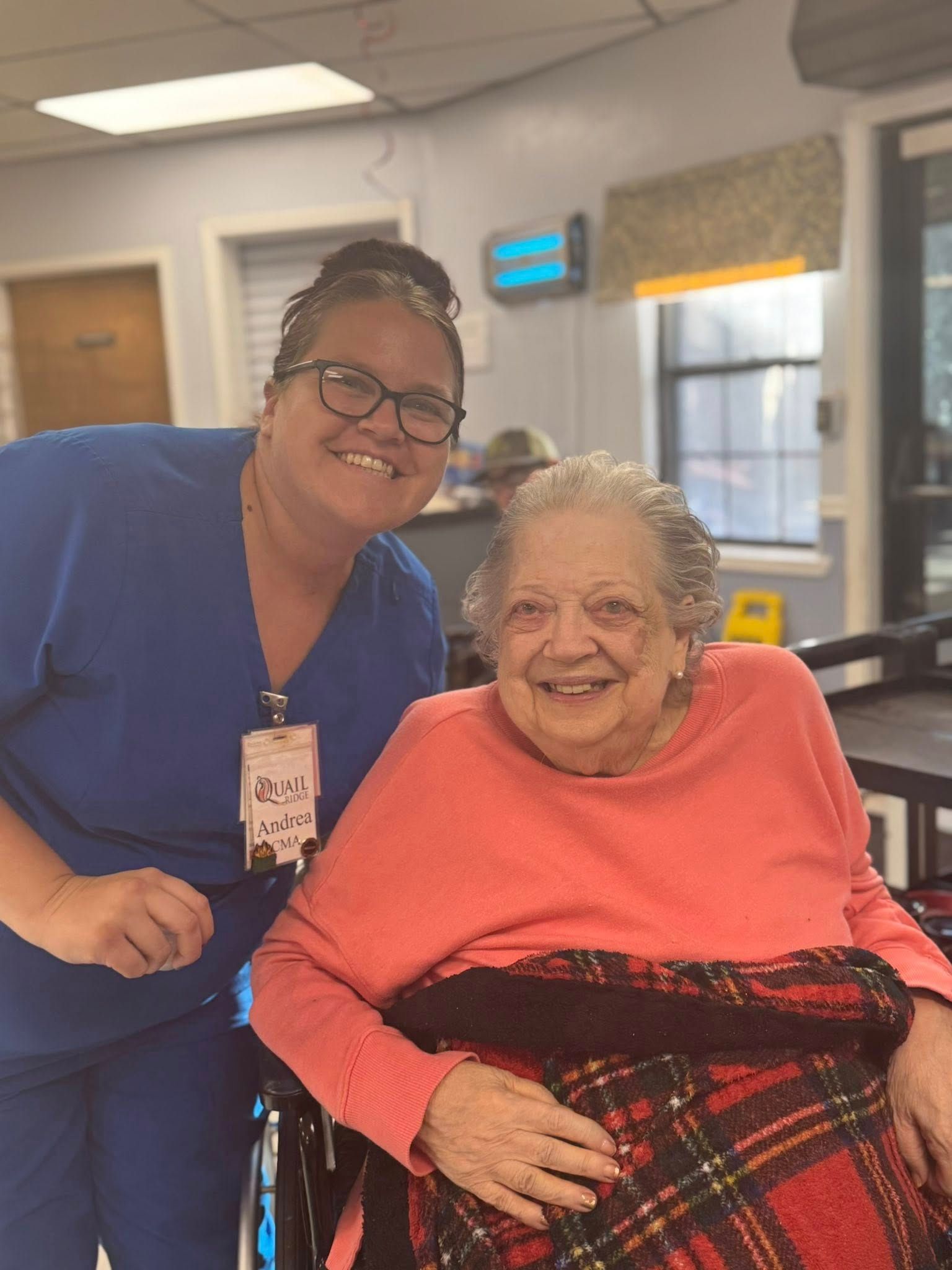 Caregiver and elderly woman in a wheelchair smiling together. Inside, caregiver in blue scrubs, elderly woman in red.