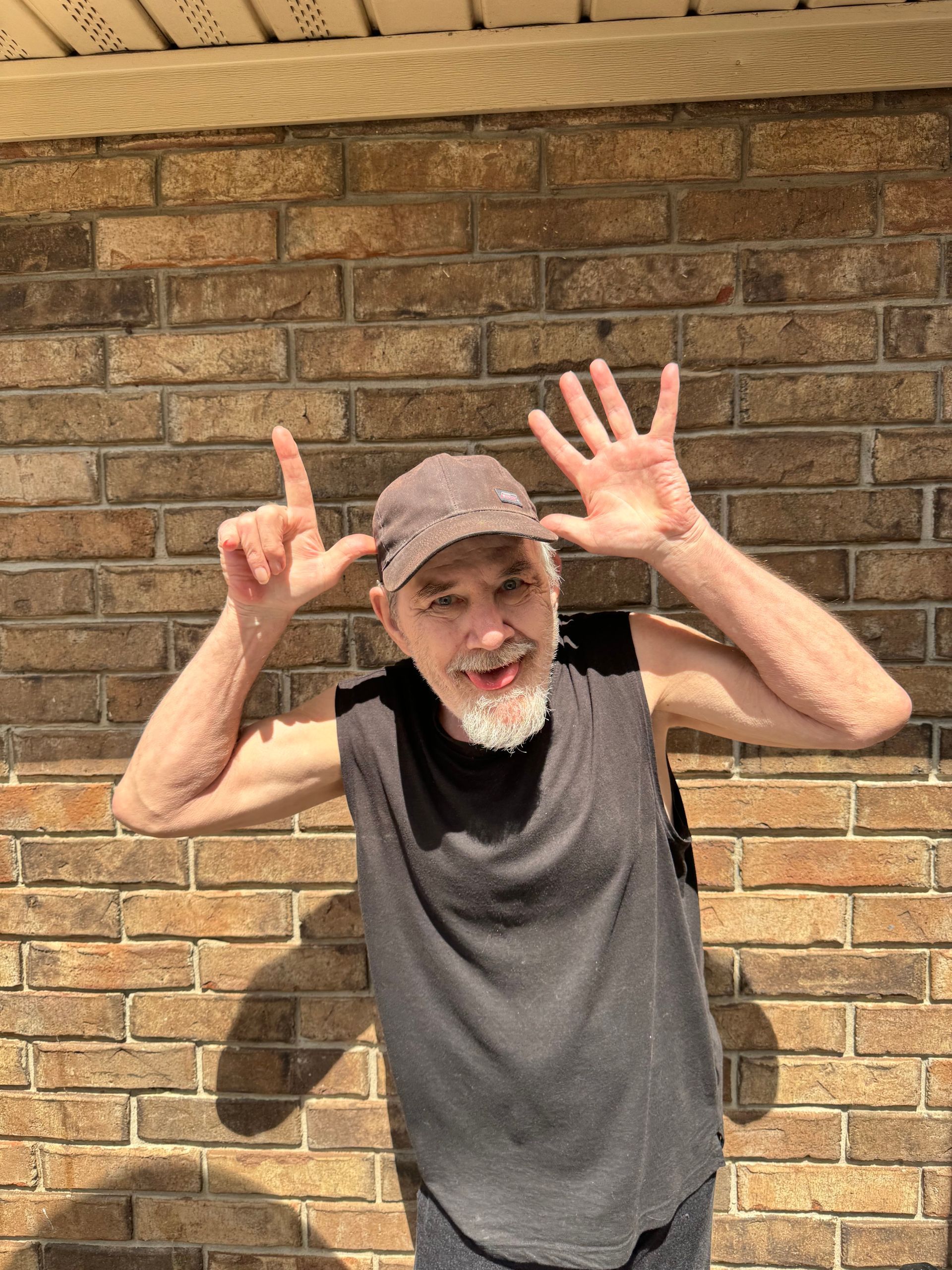 Man in dark shirt and cap, smiling and holding up 7 fingers in front of a brick wall.