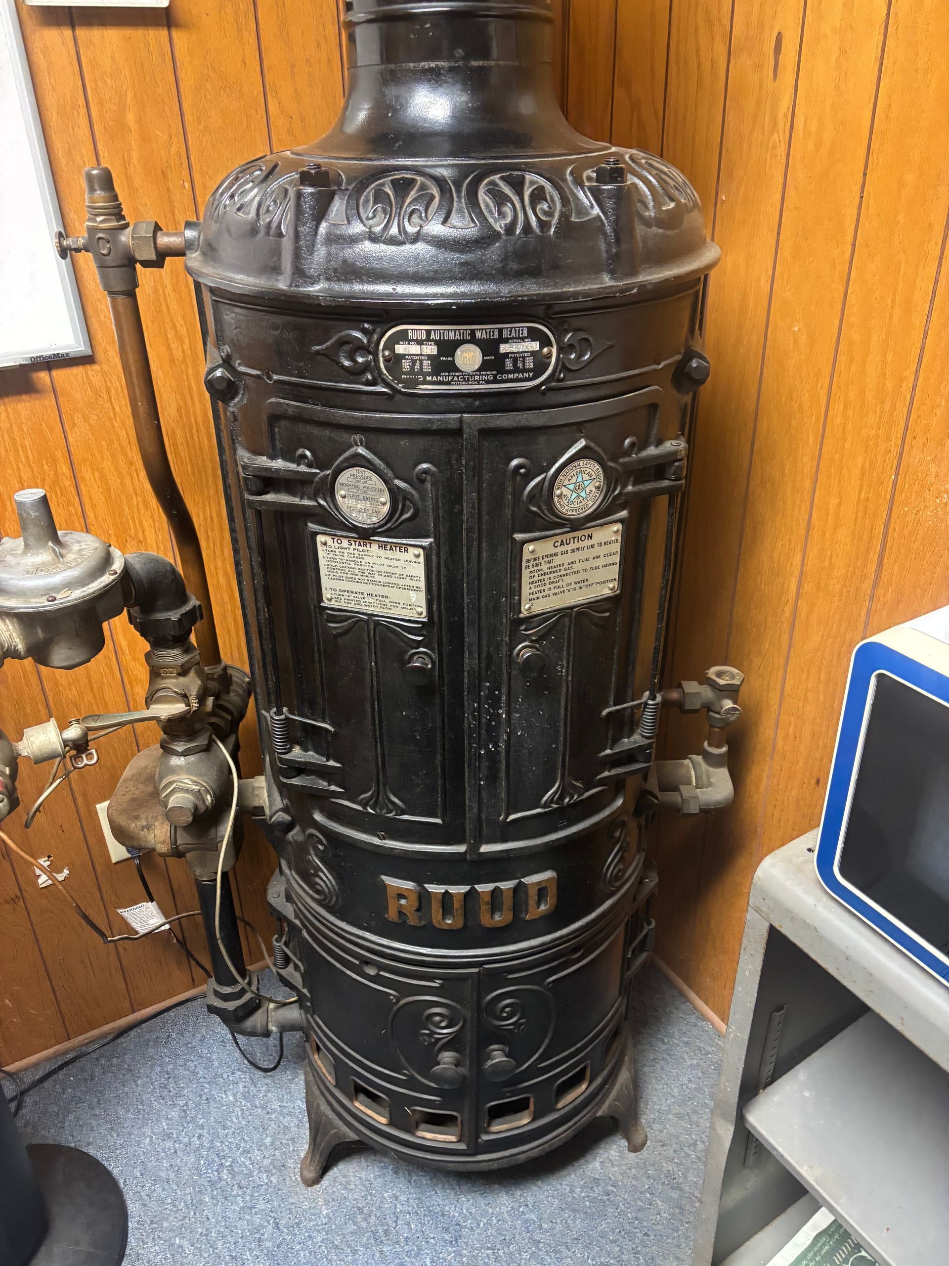 Black vintage RUUD boiler in a wood-paneled room, with exposed pipes and a small monitor nearby Black vintage RUUD boiler in a wood-paneled room, with exposed pipes and a small monitor nearby