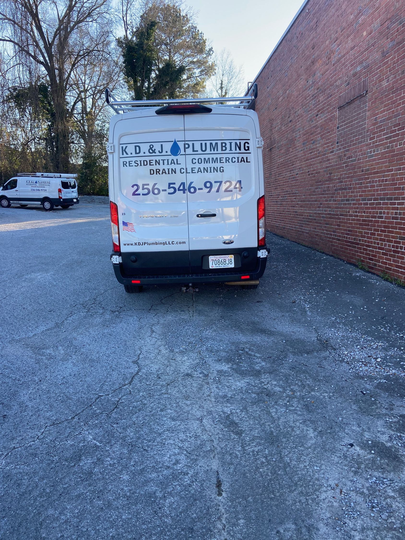 White work van parked beside a brick building on a gravel lot, with trees and another vehicle nearby. White work van parked beside a brick building on a gravel lot, with trees and another vehicle nearby.