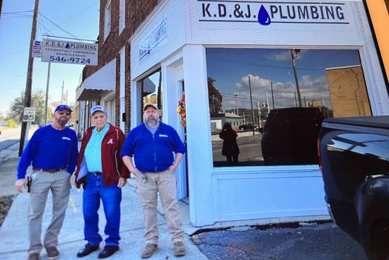Three men stand outside K.D.&J. Plumbing storefront on a sunny street. Three men stand outside K.D.&J. Plumbing storefront on a sunny street.