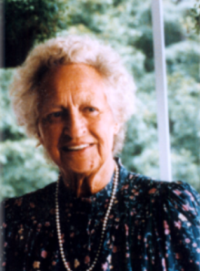 An elderly woman wearing a pearl necklace smiles for the camera