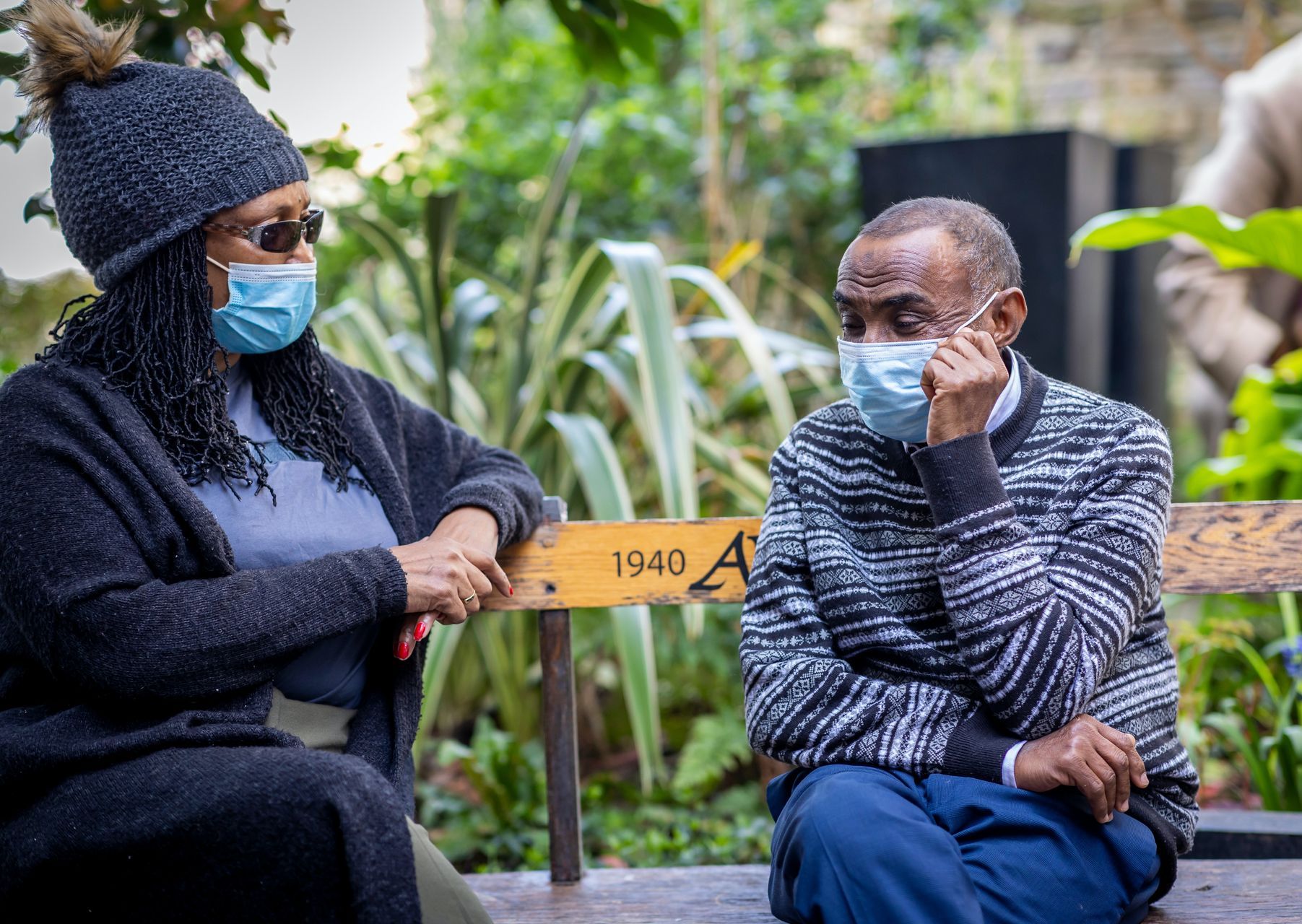 A man and a woman wearing face masks are sitting on a bench.
