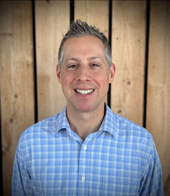 Man in blue plaid shirt smiles in front of a wooden background. Travel Advisor Franklin,TN