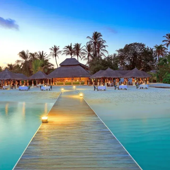 Wooden pier leading to beachside restaurant, lit at dusk. Turquoise water, palm trees, and clear sky.