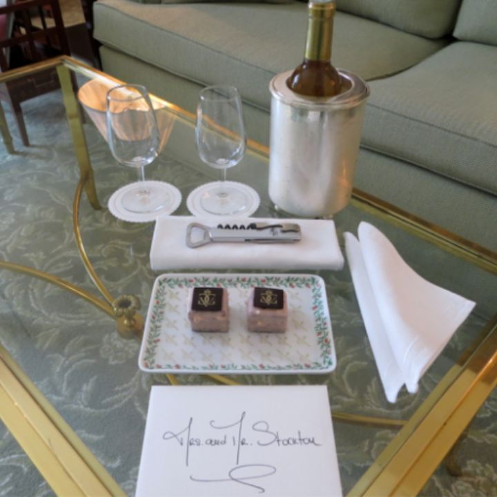 Wine bottle, glasses, chocolates, and note on a glass table in a living room.