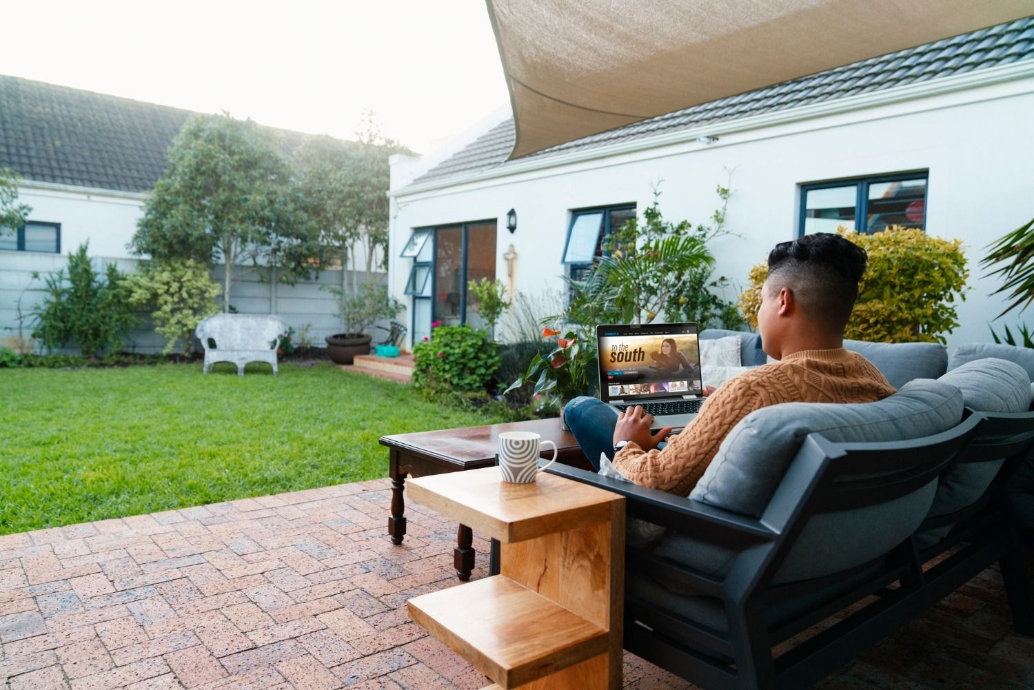 Man on outdoor sofa using laptop, looking at the screen. Brick patio, grassy backyard.