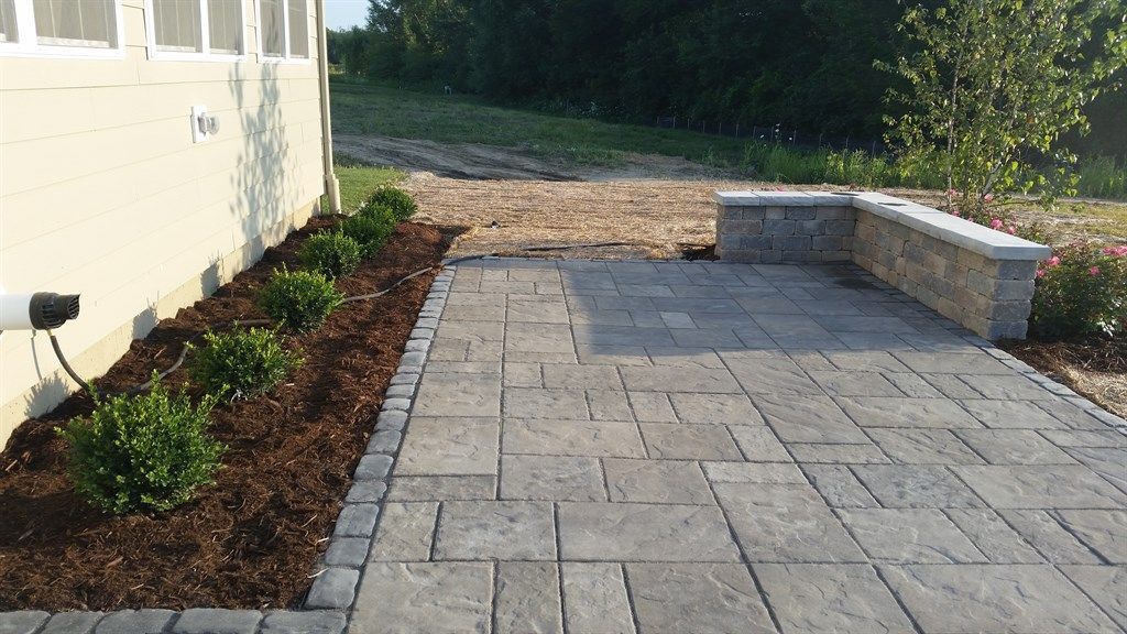 Patio with stone pavers, low wall, and shrubbery. Dark mulch lines flower bed alongside house.