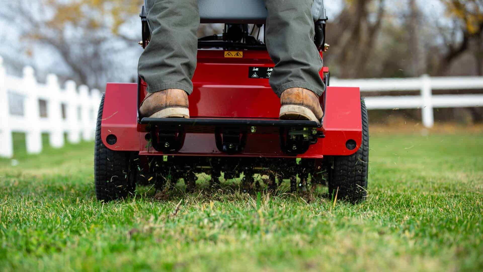 Red lawn aerator with operator, aerating a green lawn. White fence in background.