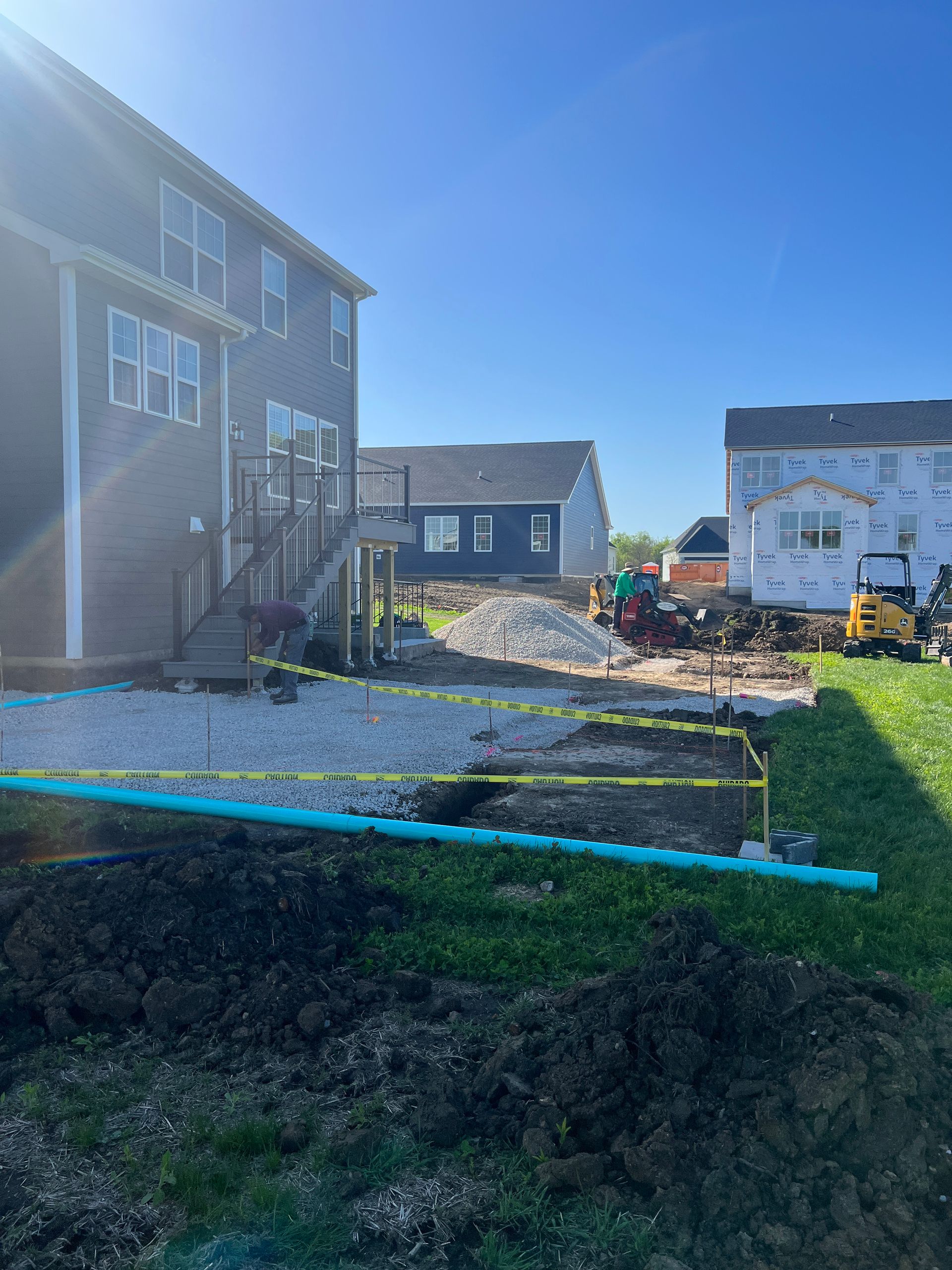 Backyard construction site; gravel, piping, and machinery near houses under a clear blue sky.