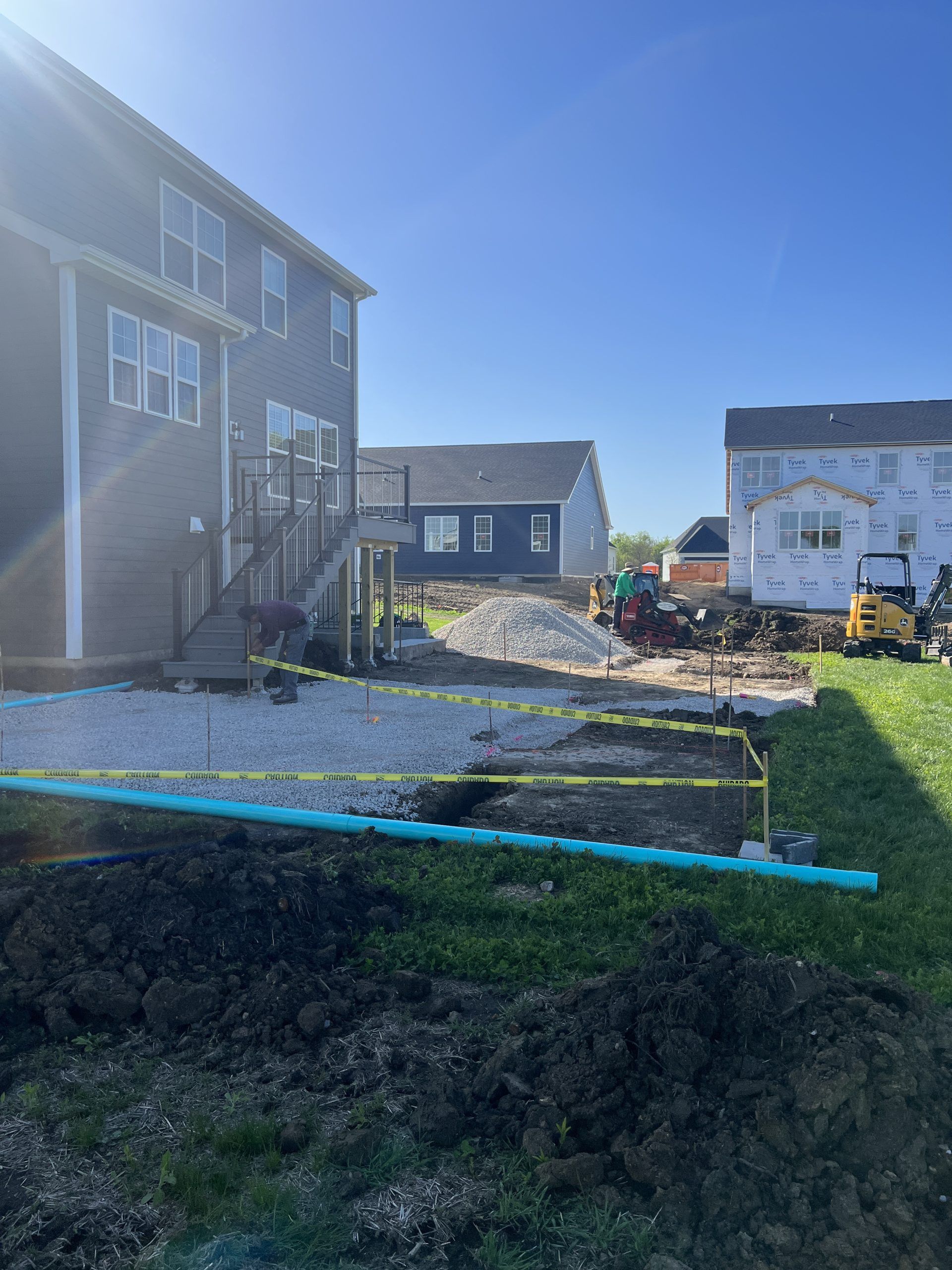 Backyard construction site; house with deck on the left, new house under construction on the right, blue pipe in foreground.