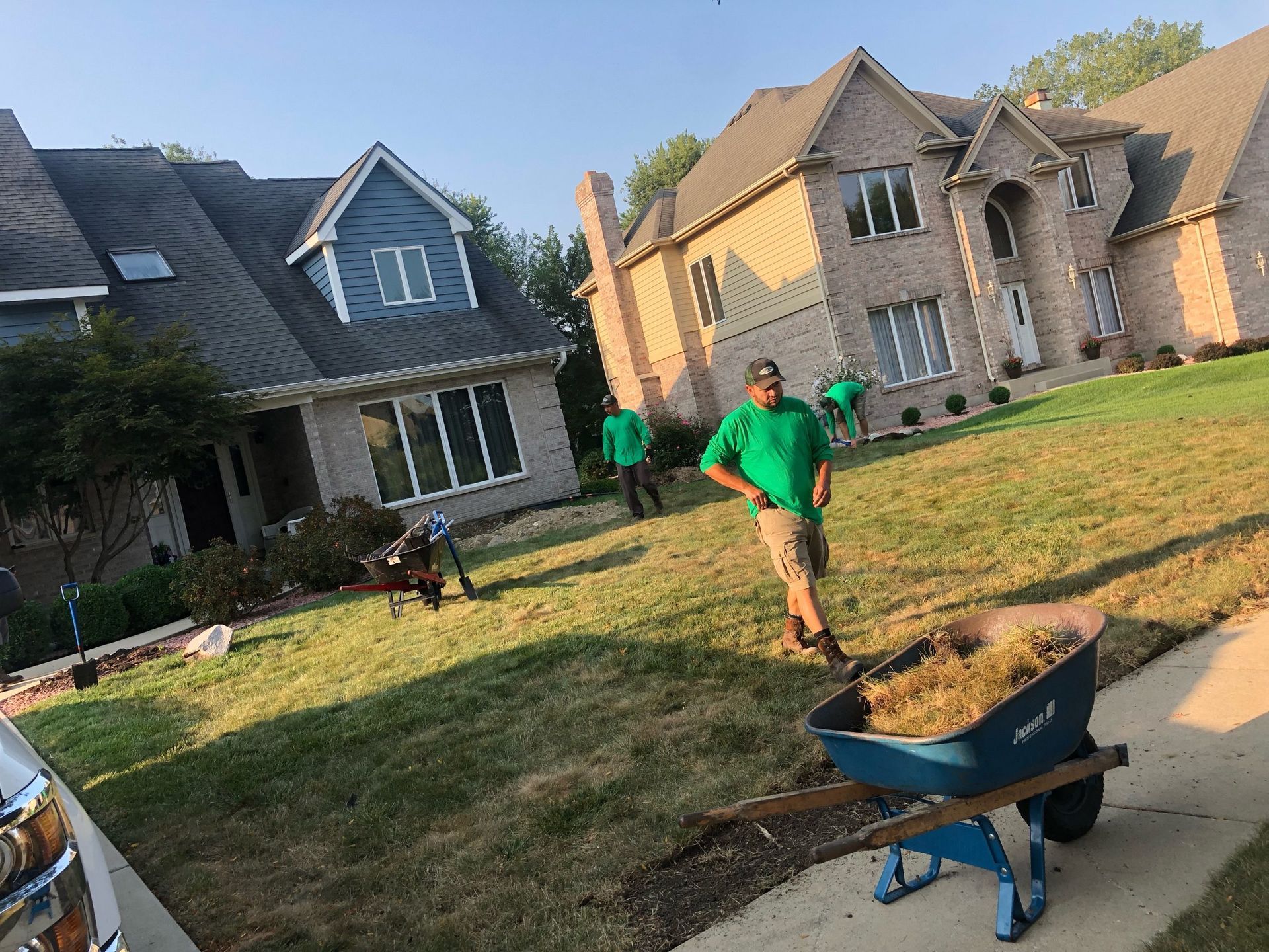 Lawn care workers mulching grass in front of two-story houses. One worker stands near a blue wheelbarrow of mulch.