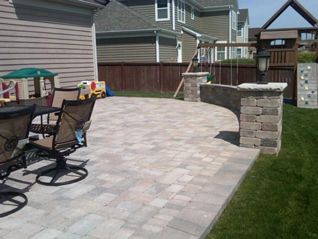 Paver patio with curved retaining wall and brick pillars near a swing set and playhouse.