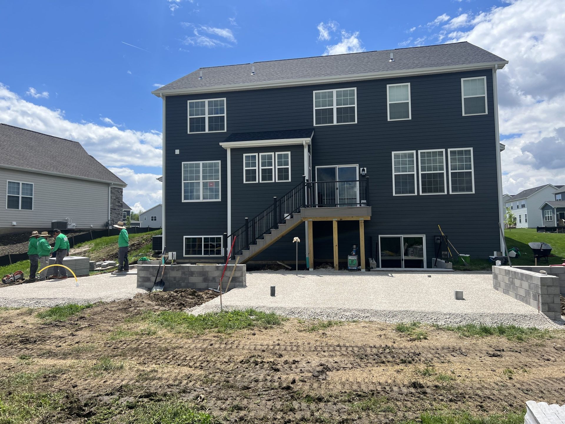 Back of a two-story blue house with deck. Construction in progress: patio with retaining walls, workers present.