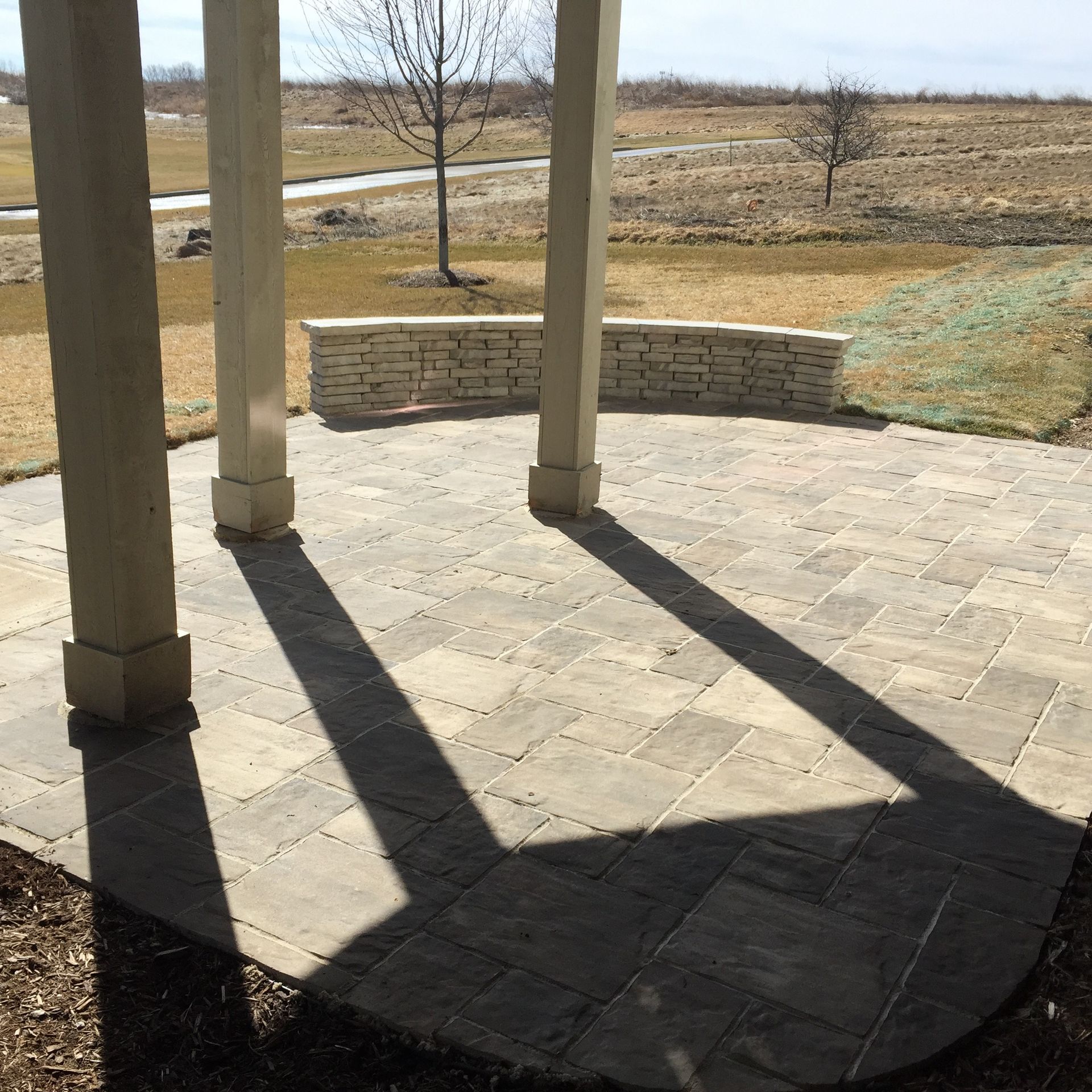 Patio with three columns casting shadows on paved floor, low stone wall, and grassy field.