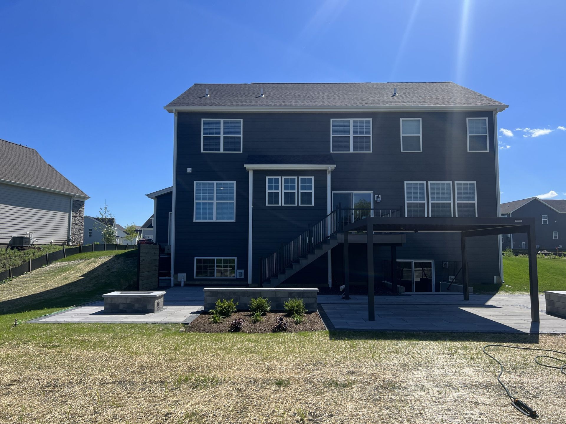 Back of a two-story dark blue house with patio, pergola, and fire pit. Blue sky.