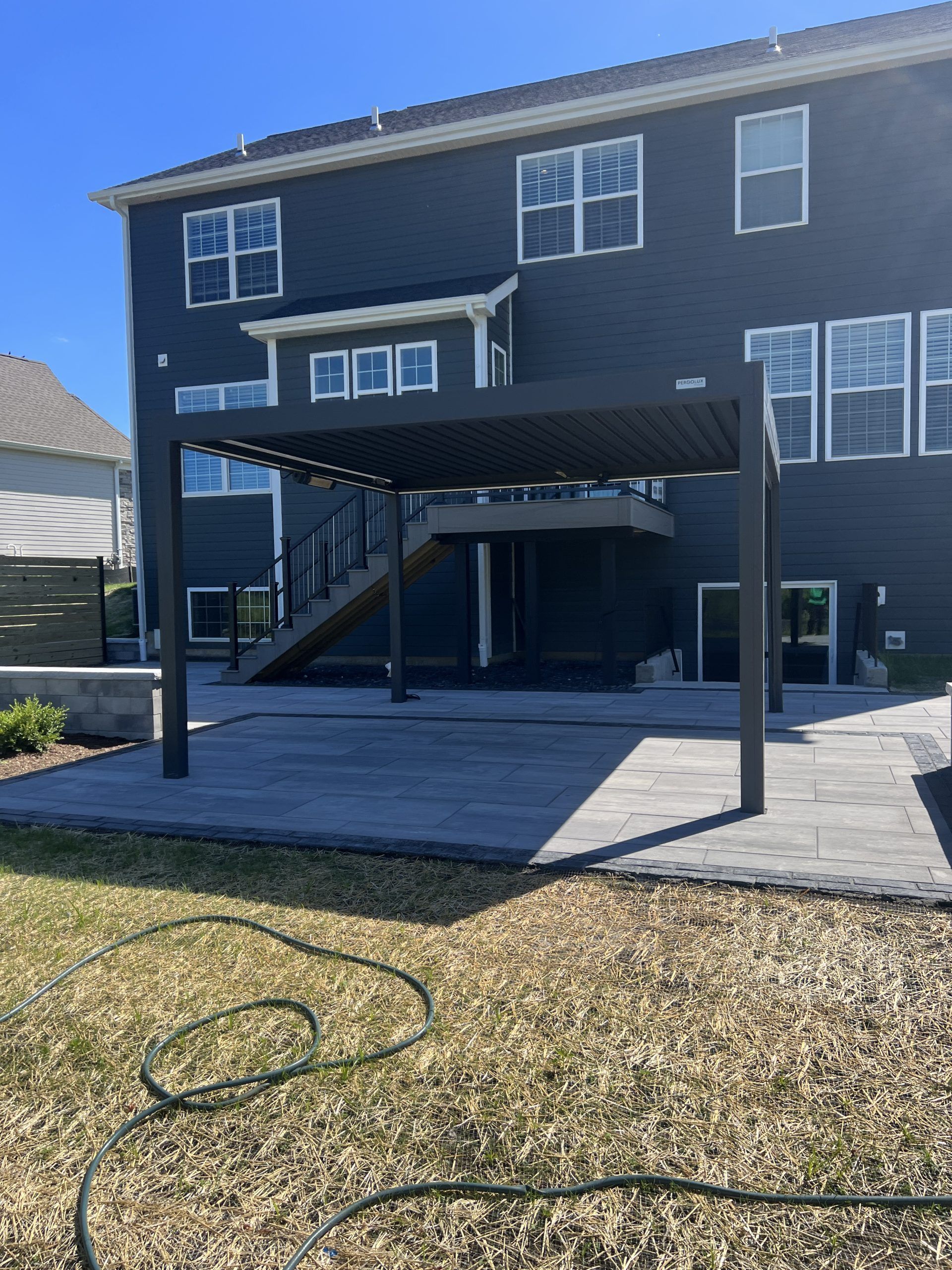 Black pergola on a gray patio with a dark blue house in the background. Sunny day.