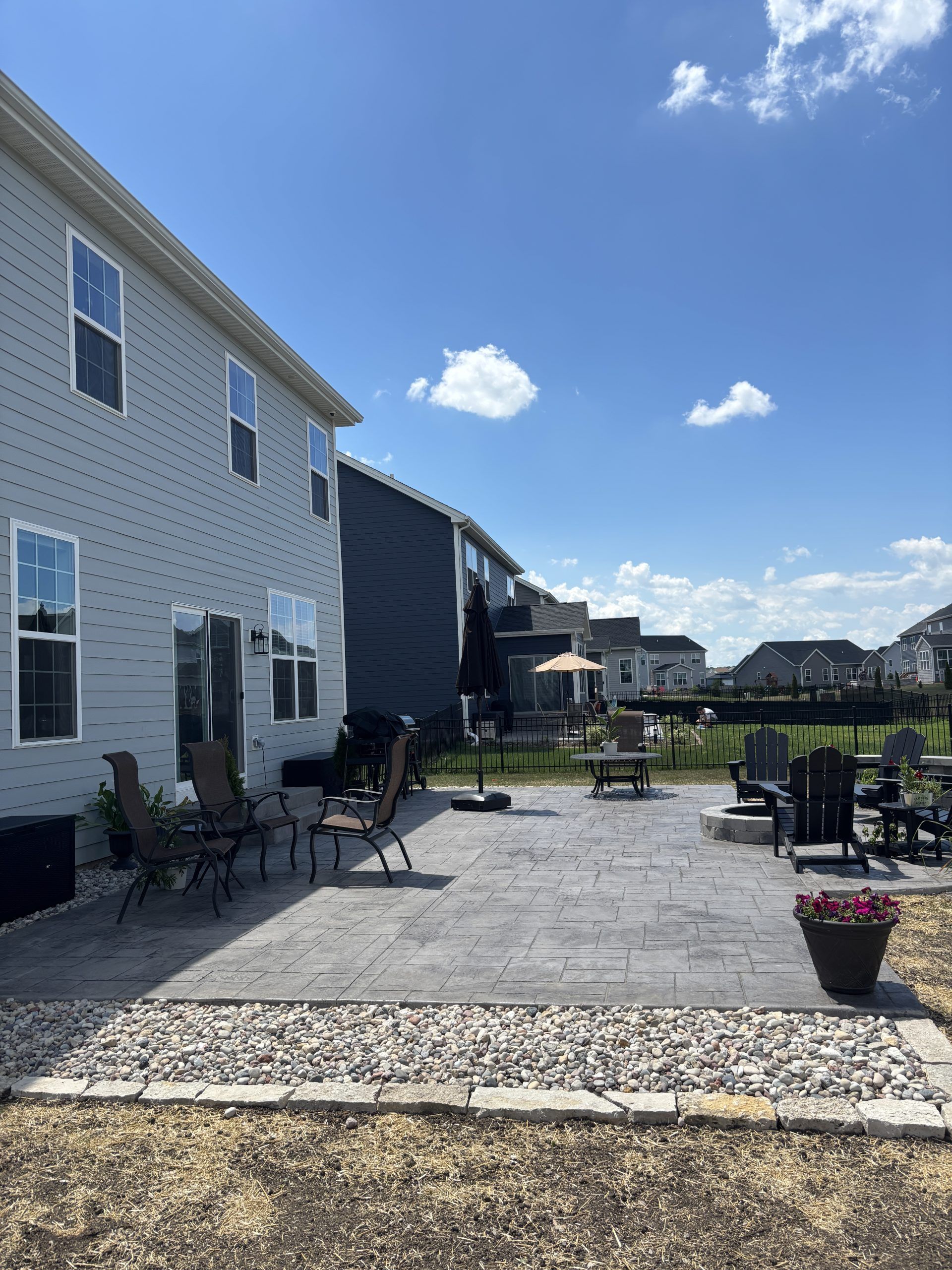 Backyard patio with chairs, umbrella, and a grill, next to a two-story gray house under a bright blue sky.