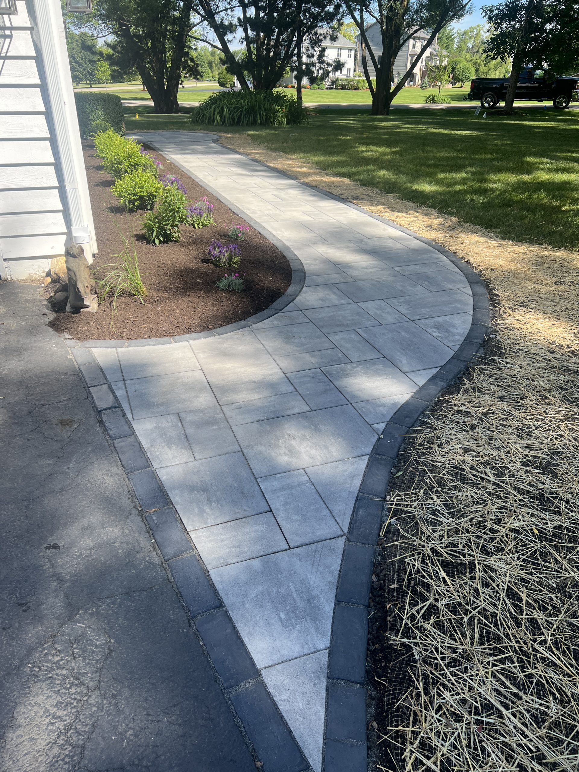 A curving paved walkway with dark border, next to a building and garden bed.