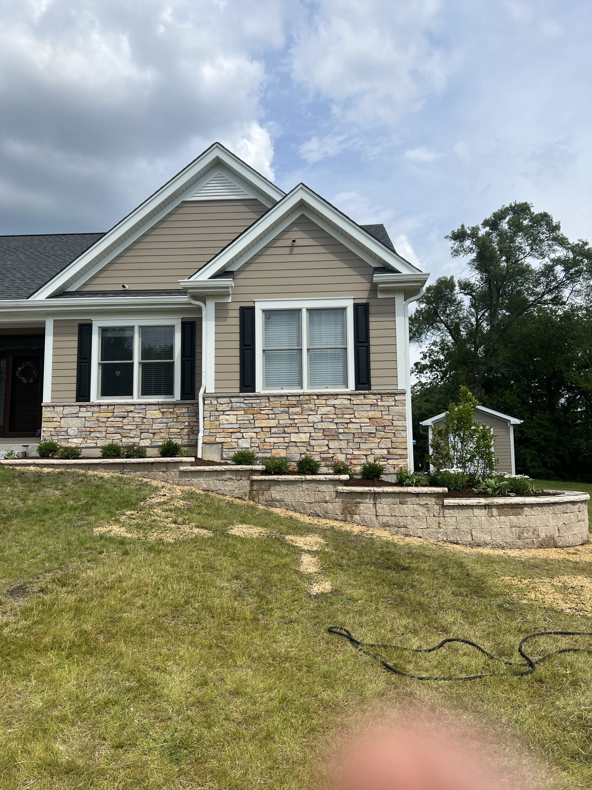 Beige house with stone accents, black shutters, and landscaping on a grassy hill under a cloudy sky.