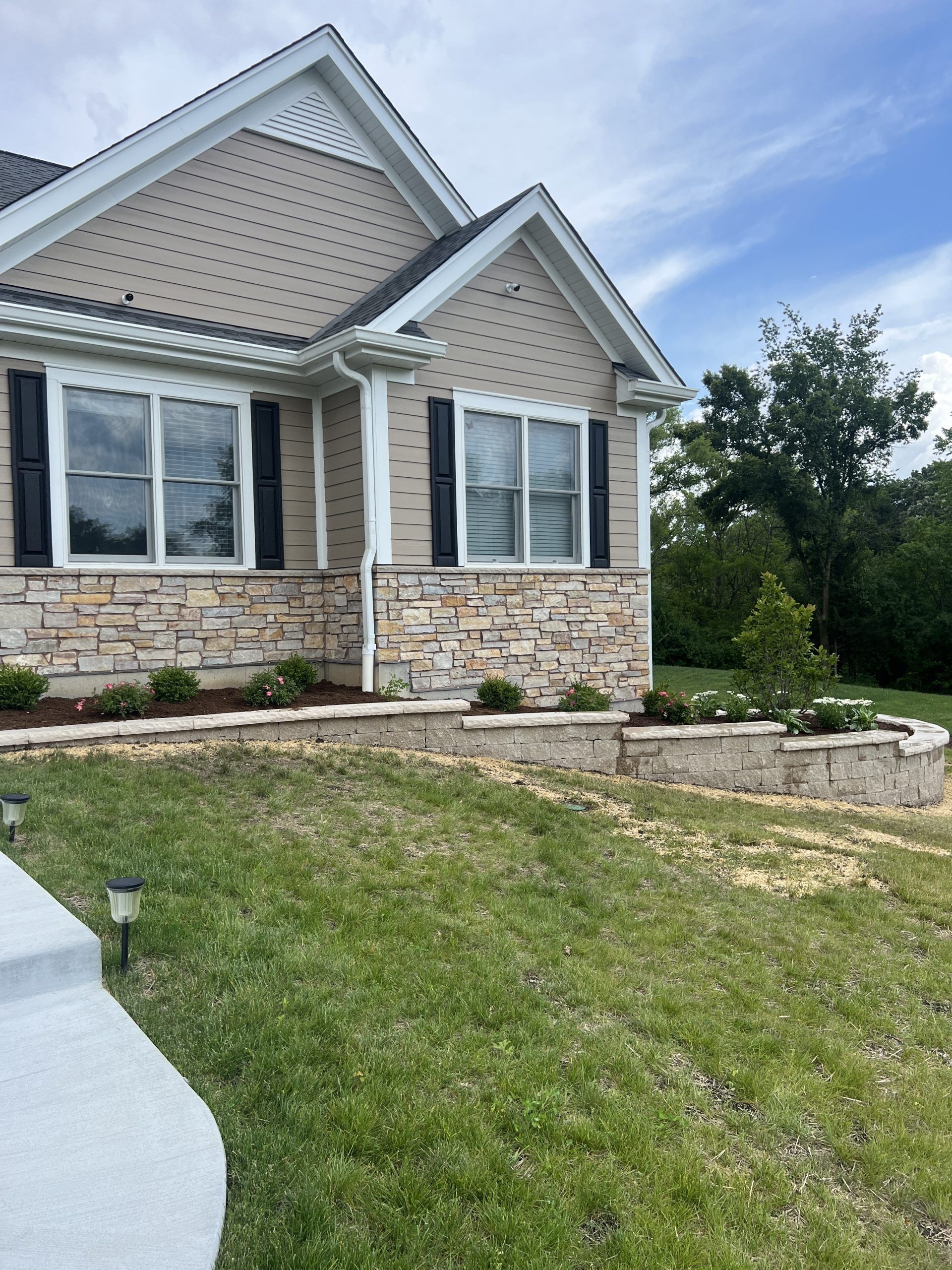Tan and stone veneer house exterior with landscaping and lawn against a partly cloudy sky.