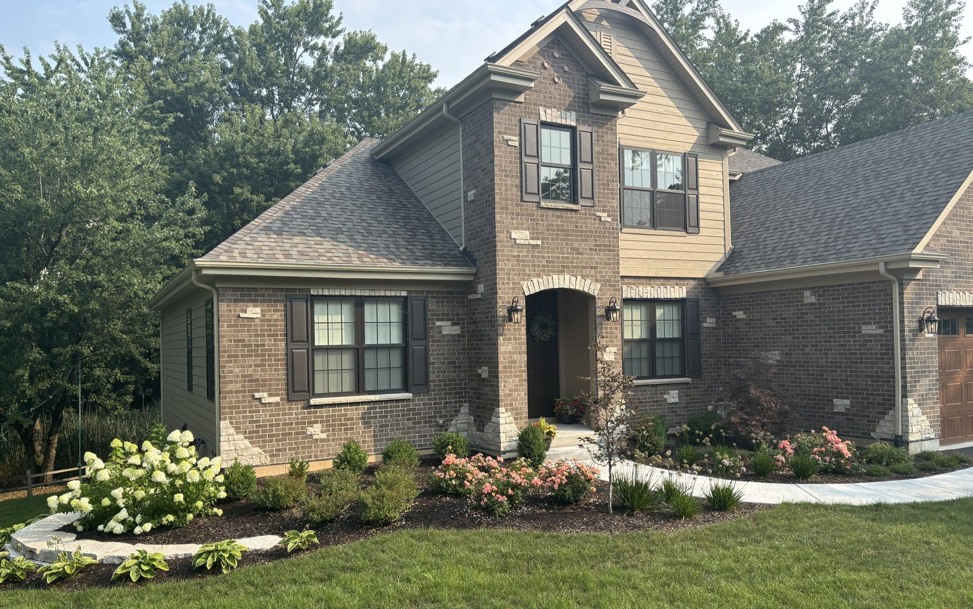 Landscaped front yard with flowering pink bushes, trees, and large stone features.