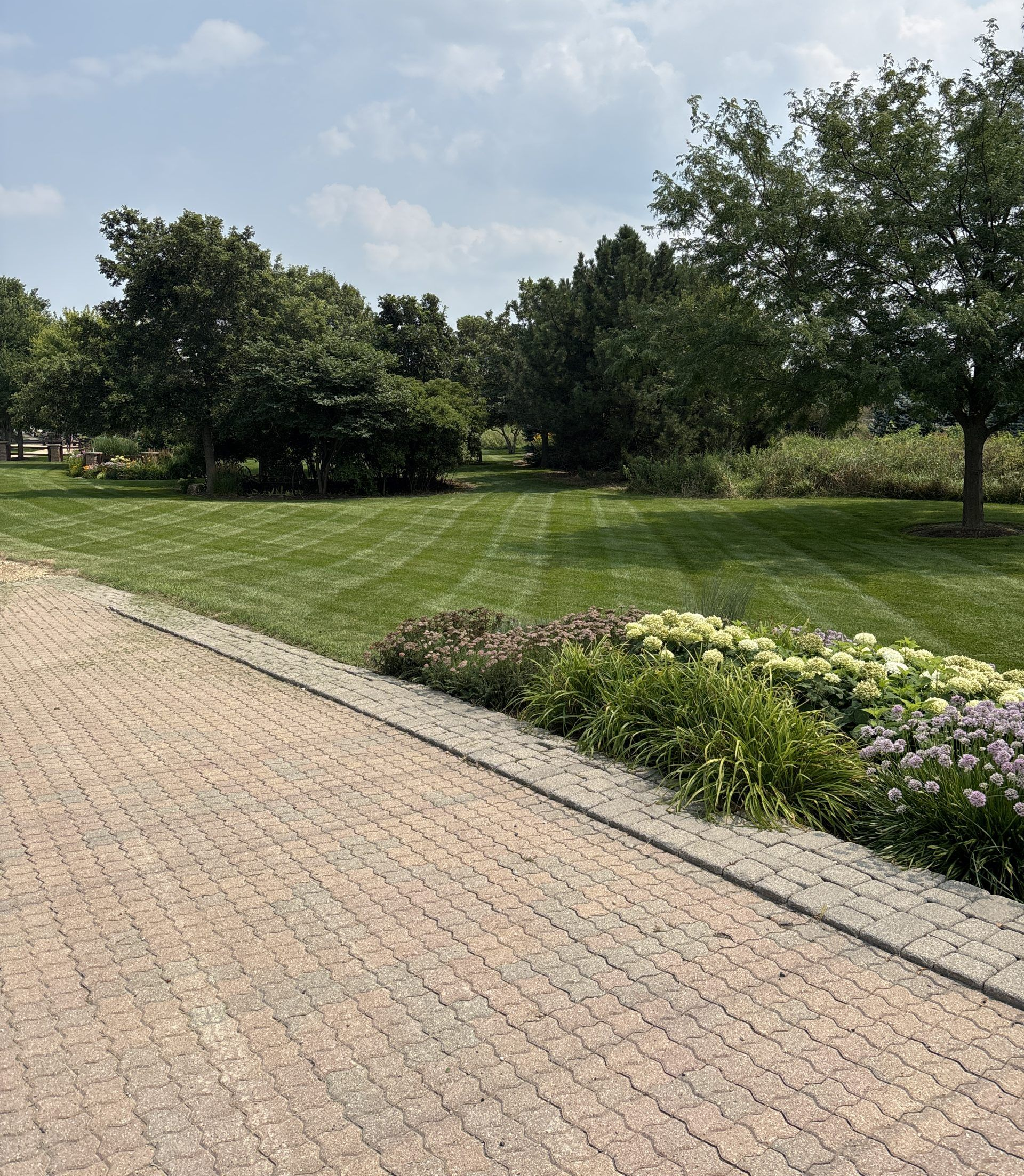 Brick path next to a manicured lawn and garden bed, trees in the background, under a blue sky.