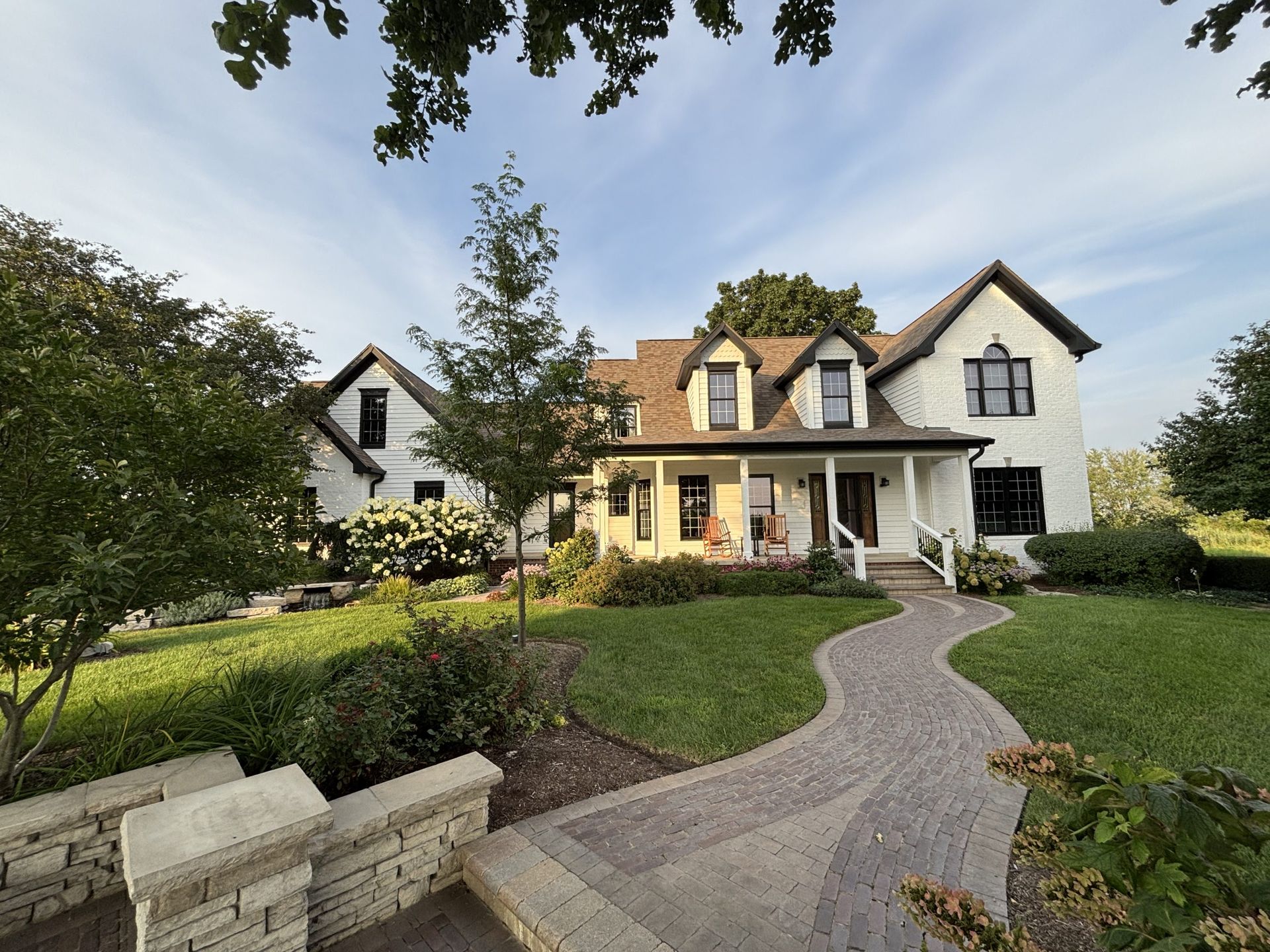 White farmhouse with black trim, surrounded by green lawn and gardens, and a stone pathway.