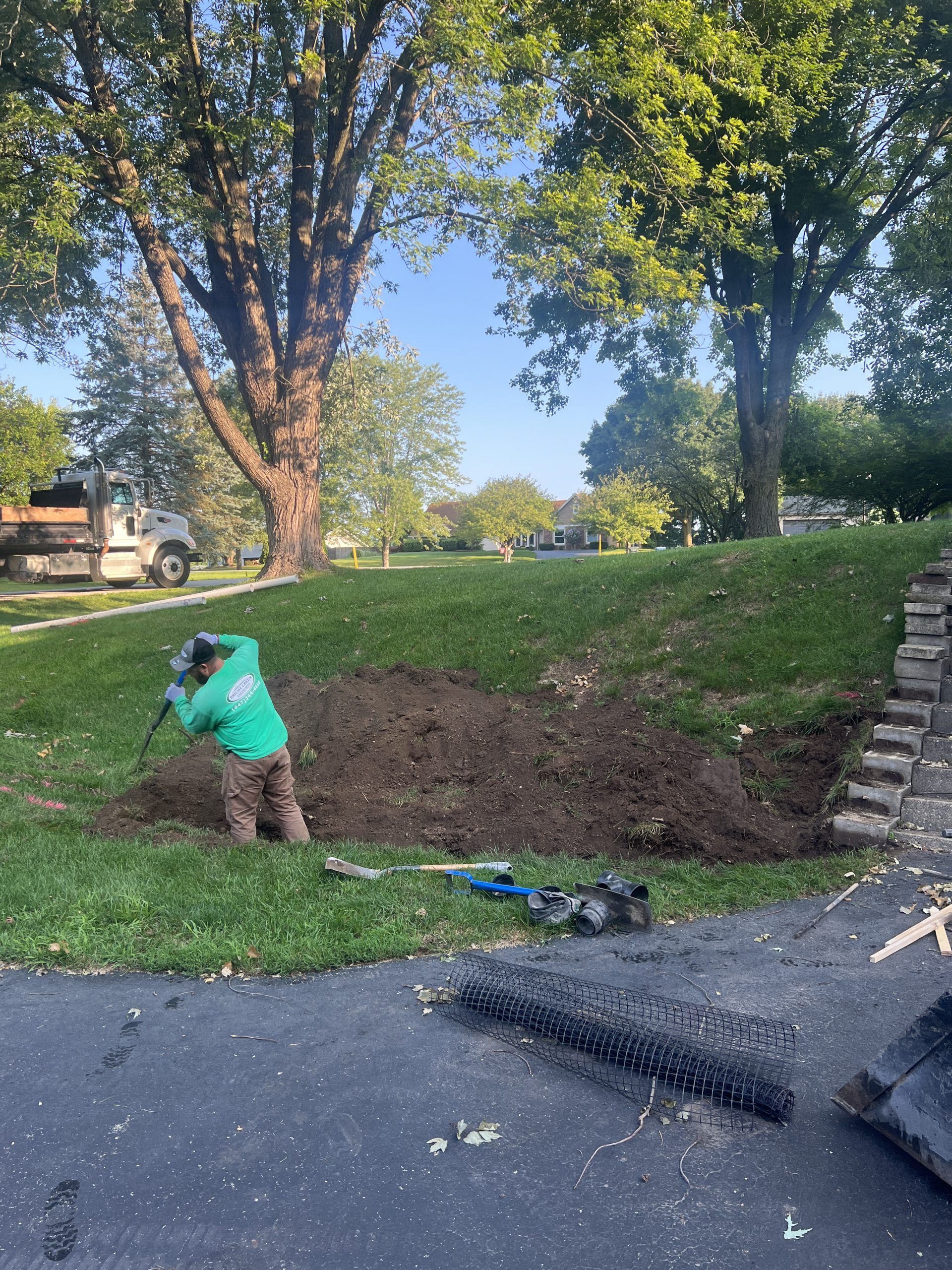 Man digging in a yard, removing soil. A truck and trees are visible in the background.