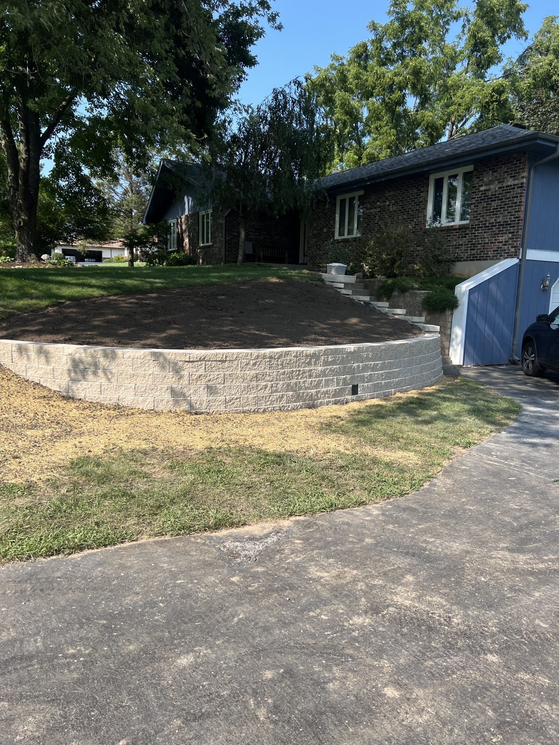 A house with a blue facade, a stone retaining wall, and a driveway on a sunny day.