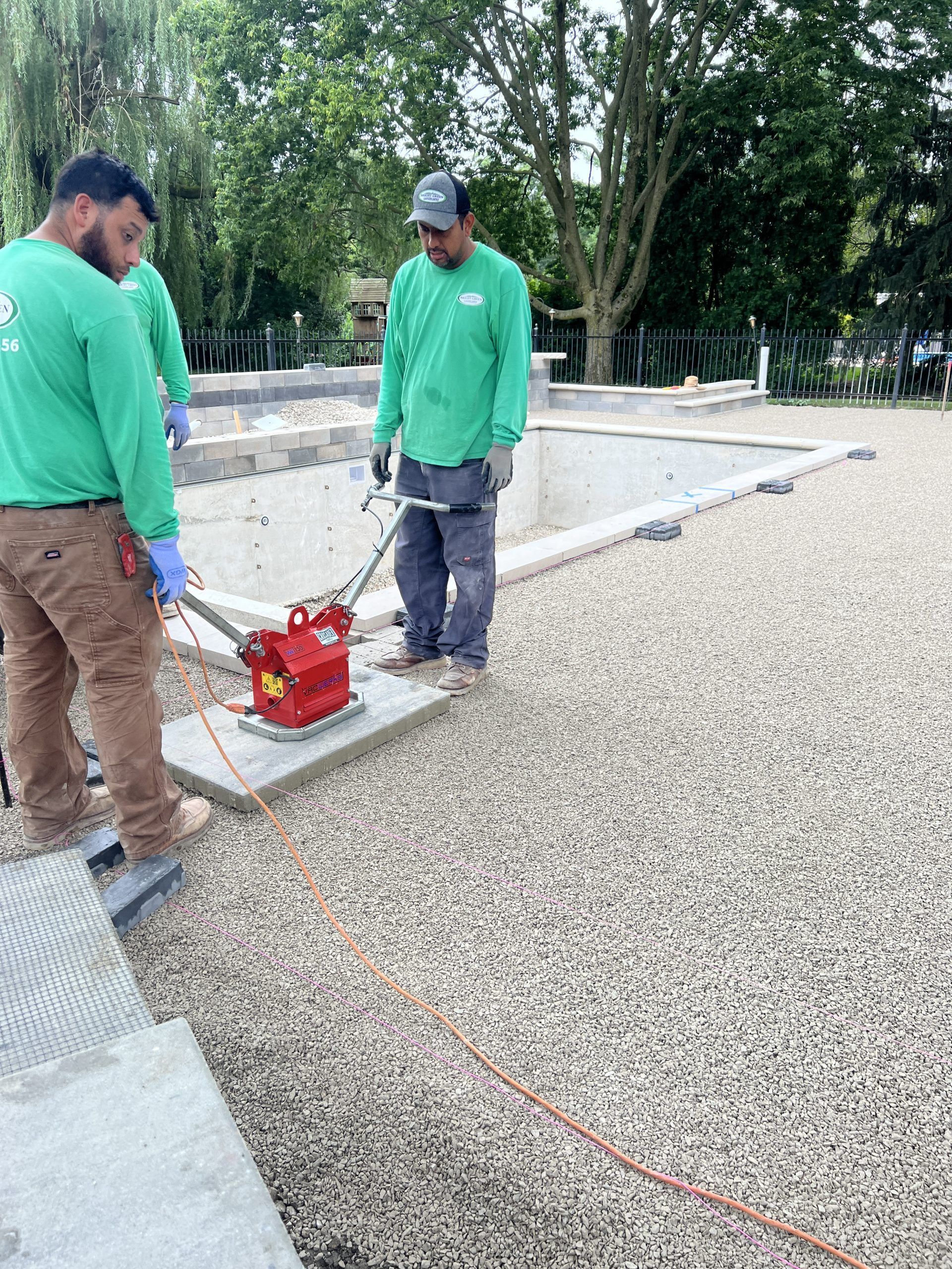 Three workers compact gravel near a pool. They wear green shirts, one operates a compactor.