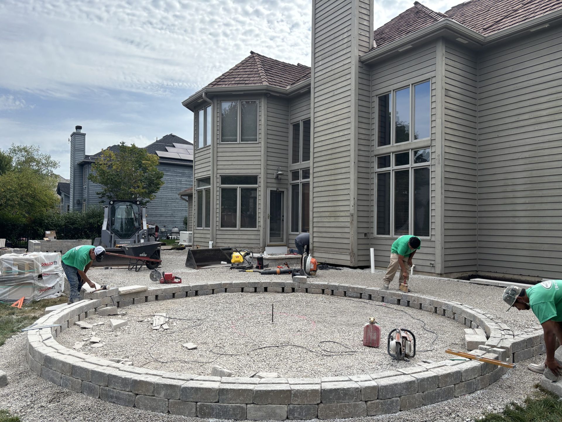 Construction workers laying pavers for a circular patio base near a large house.