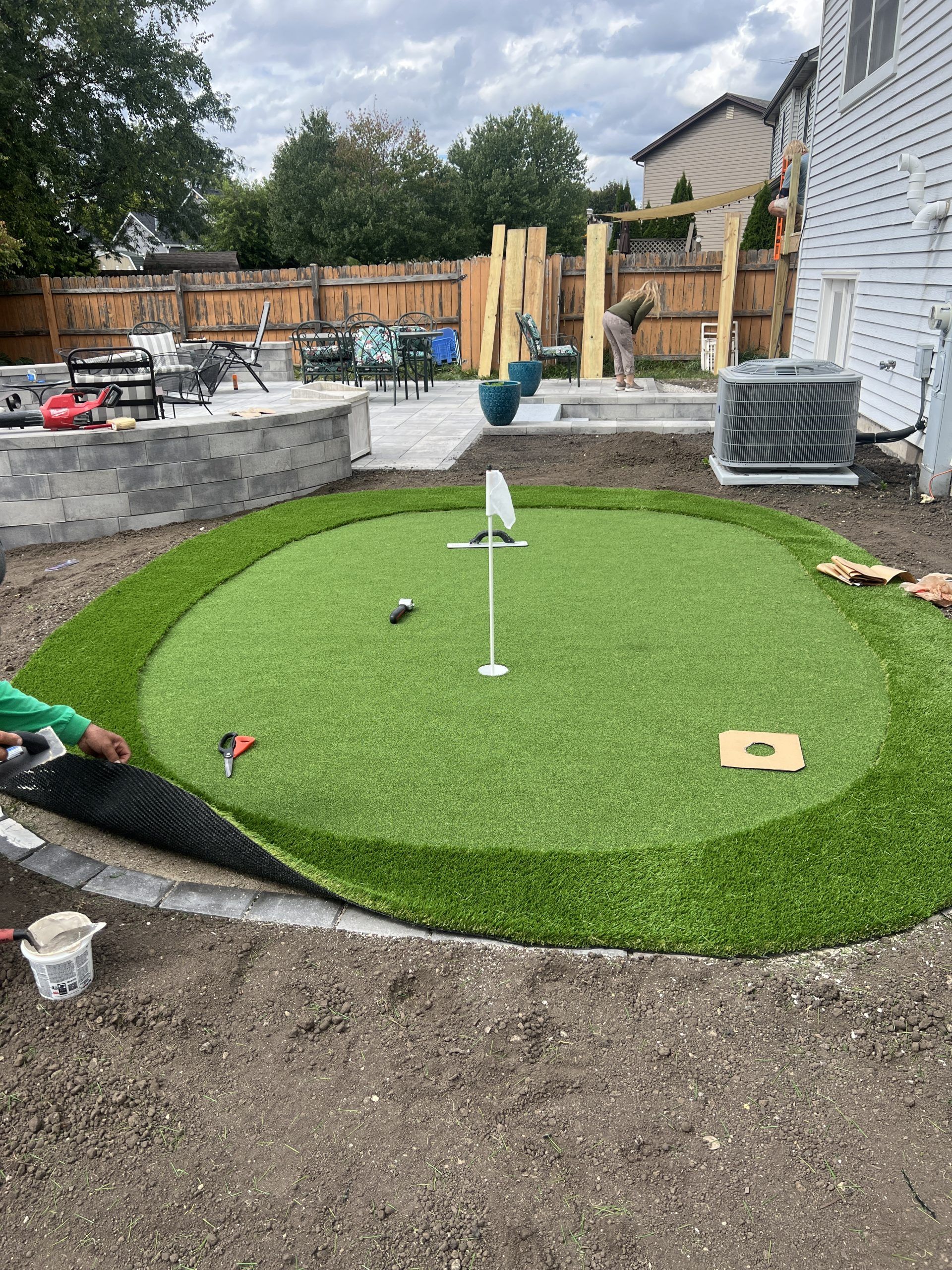 Installation of a backyard putting green, surrounded by a dark edging, with a flag and other construction supplies.