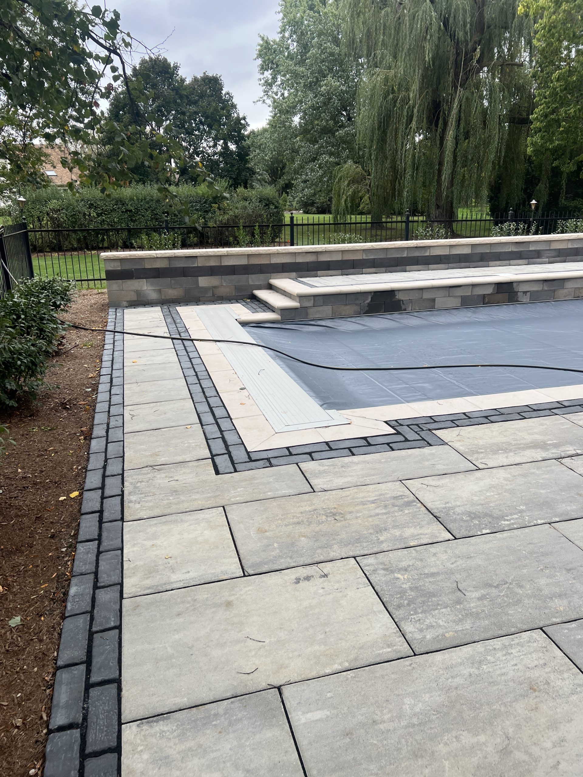 Stone patio with a dark pool cover and steps. Dark border on the patio edge. Trees in the background.