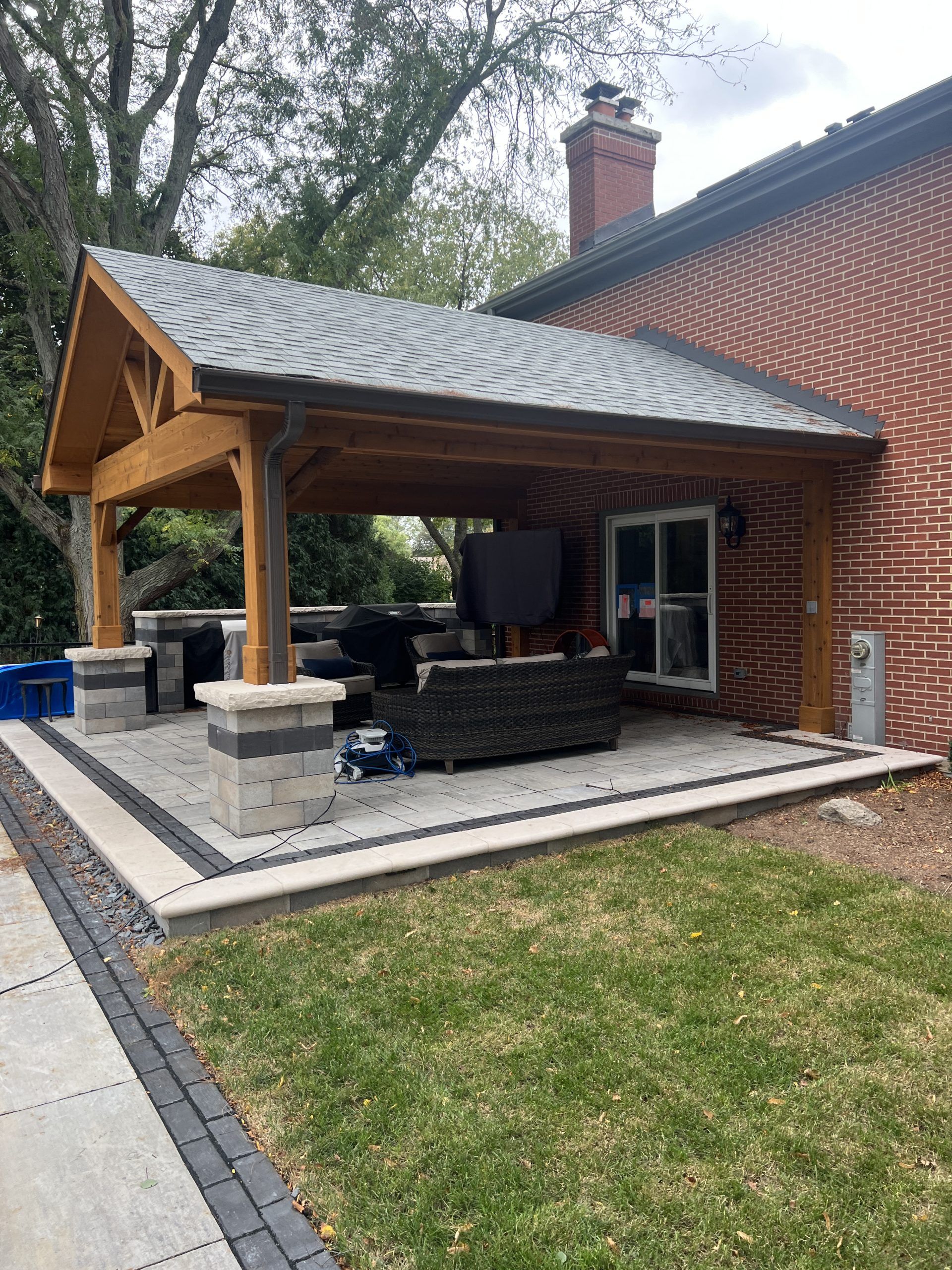 Outdoor patio with wooden roof and stone supports attached to a brick house.