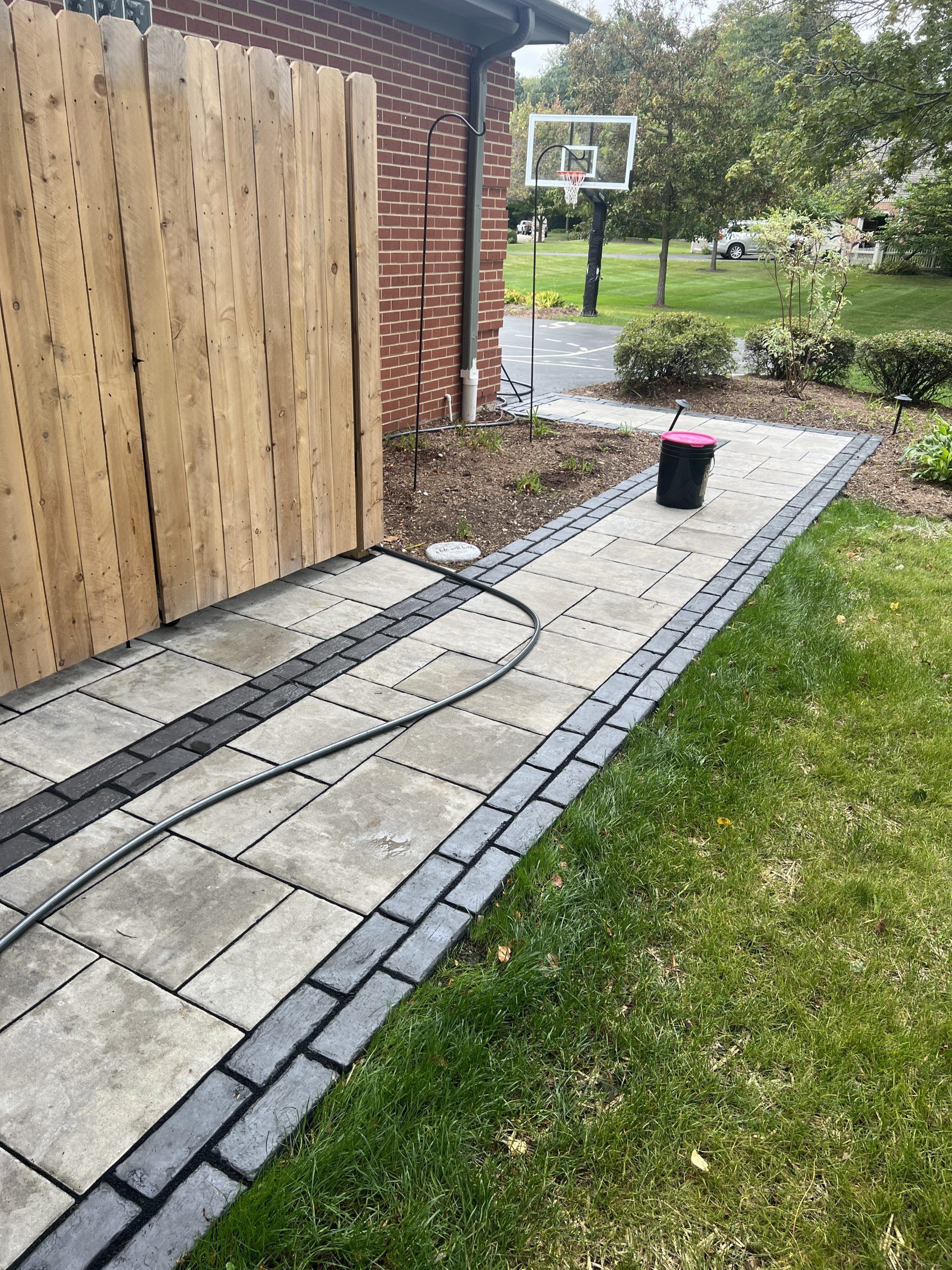 Brick walkway alongside a brick house and wooden fence. Black edging with a basketball hoop in the distance.
