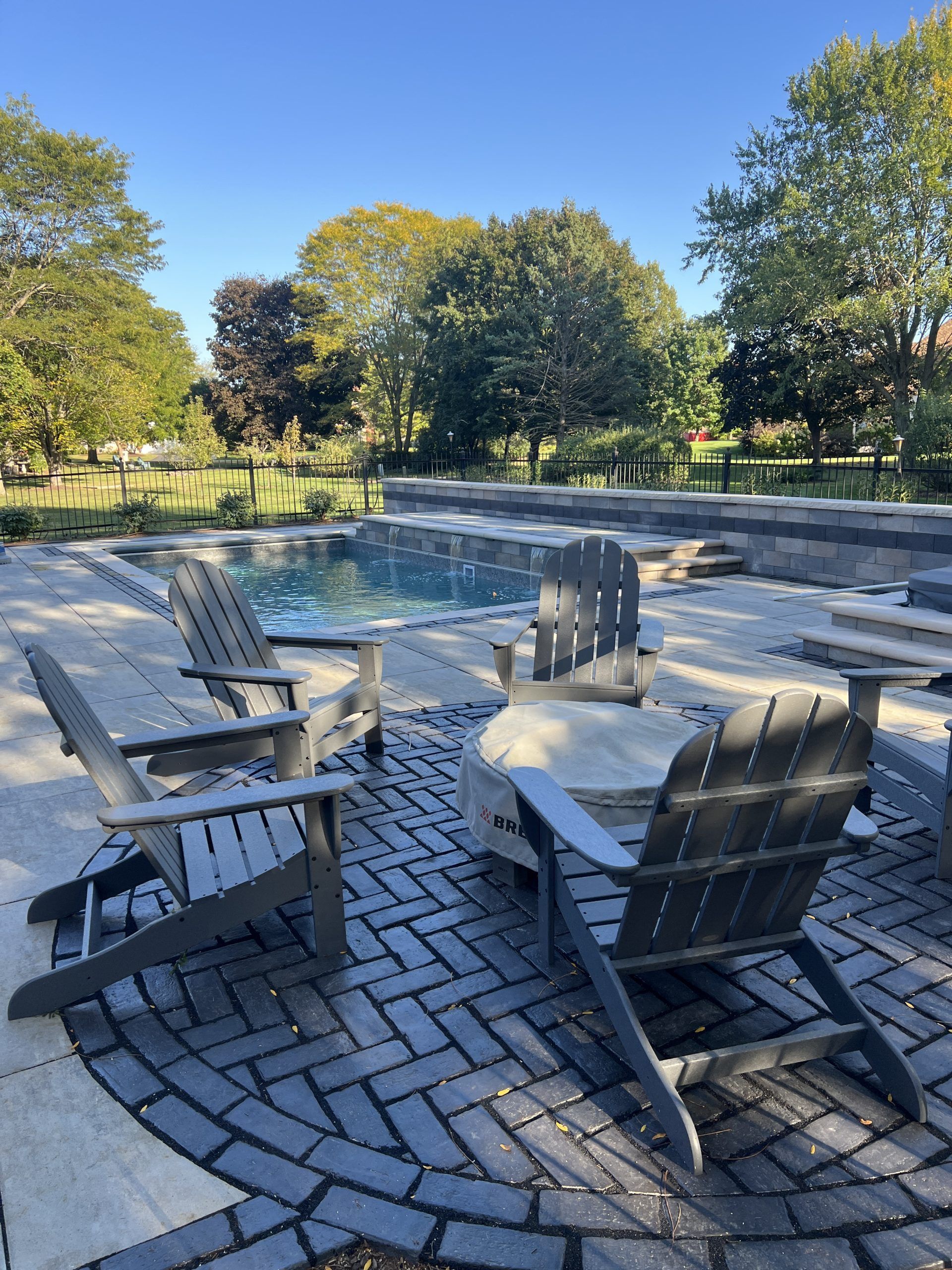 Outdoor patio with gray Adirondack chairs around a fire pit, near a pool and trees.