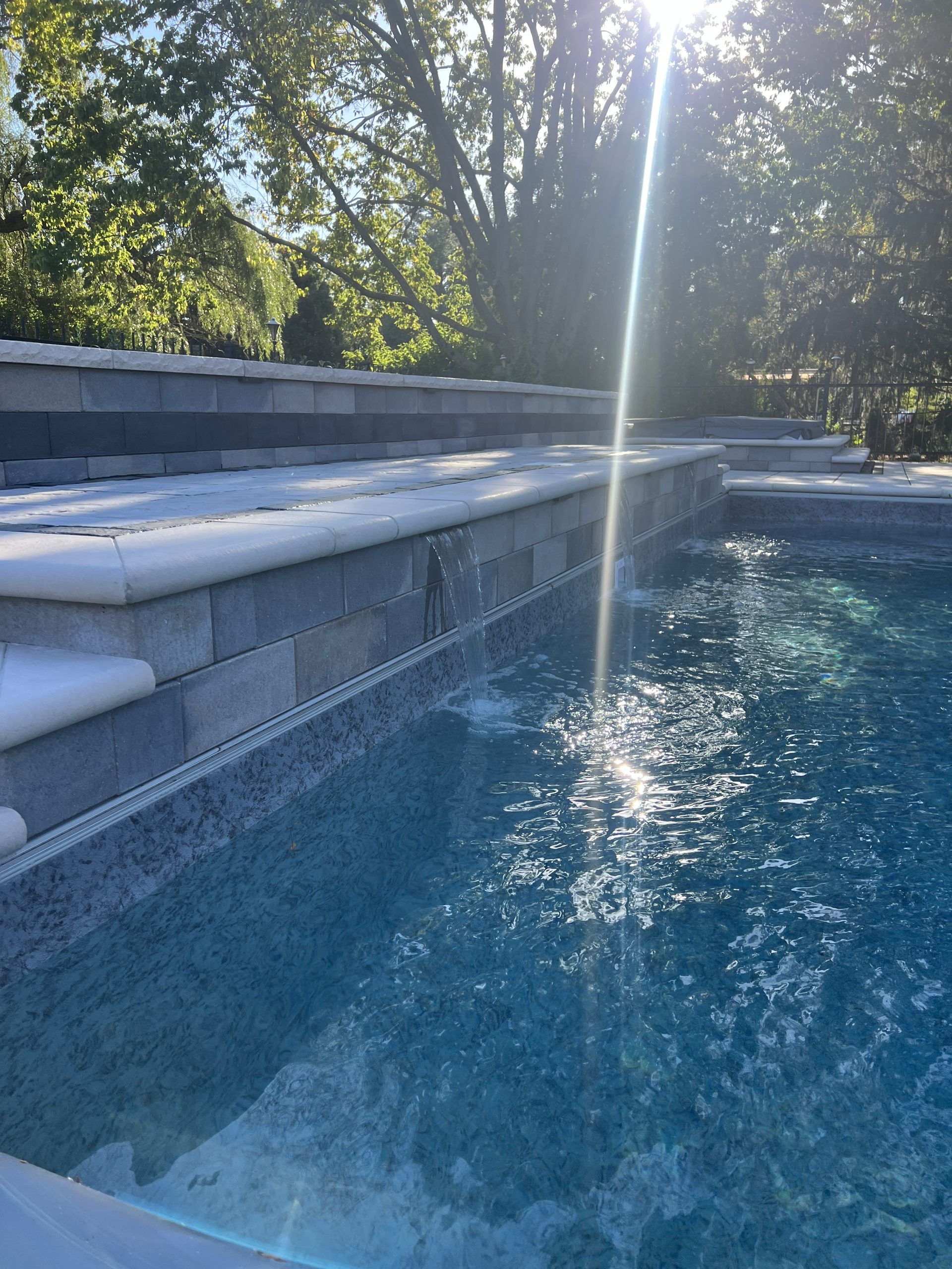 Pool with stone steps, water flowing over edge. Sun shining brightly.