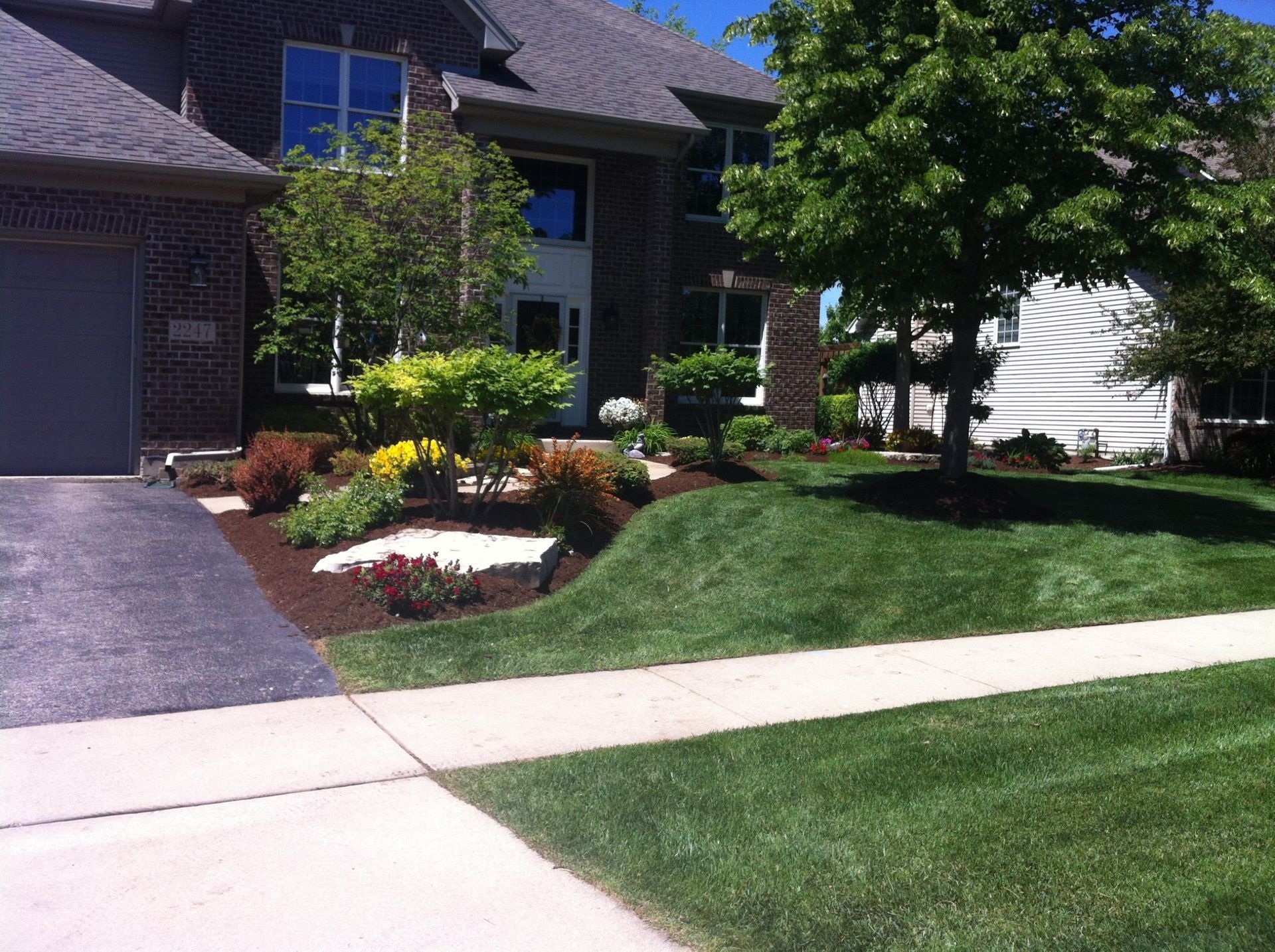 House with well-manicured lawn and landscaping. Driveway leads to brick home, sunny day.