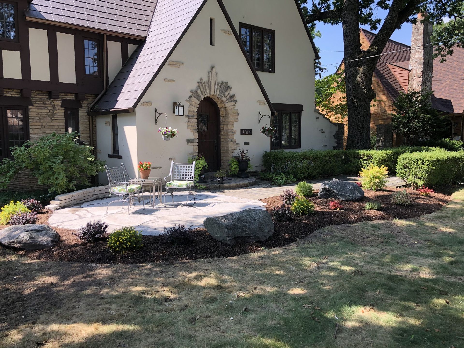 Stone patio with chairs in front of a Tudor-style house with manicured landscaping.