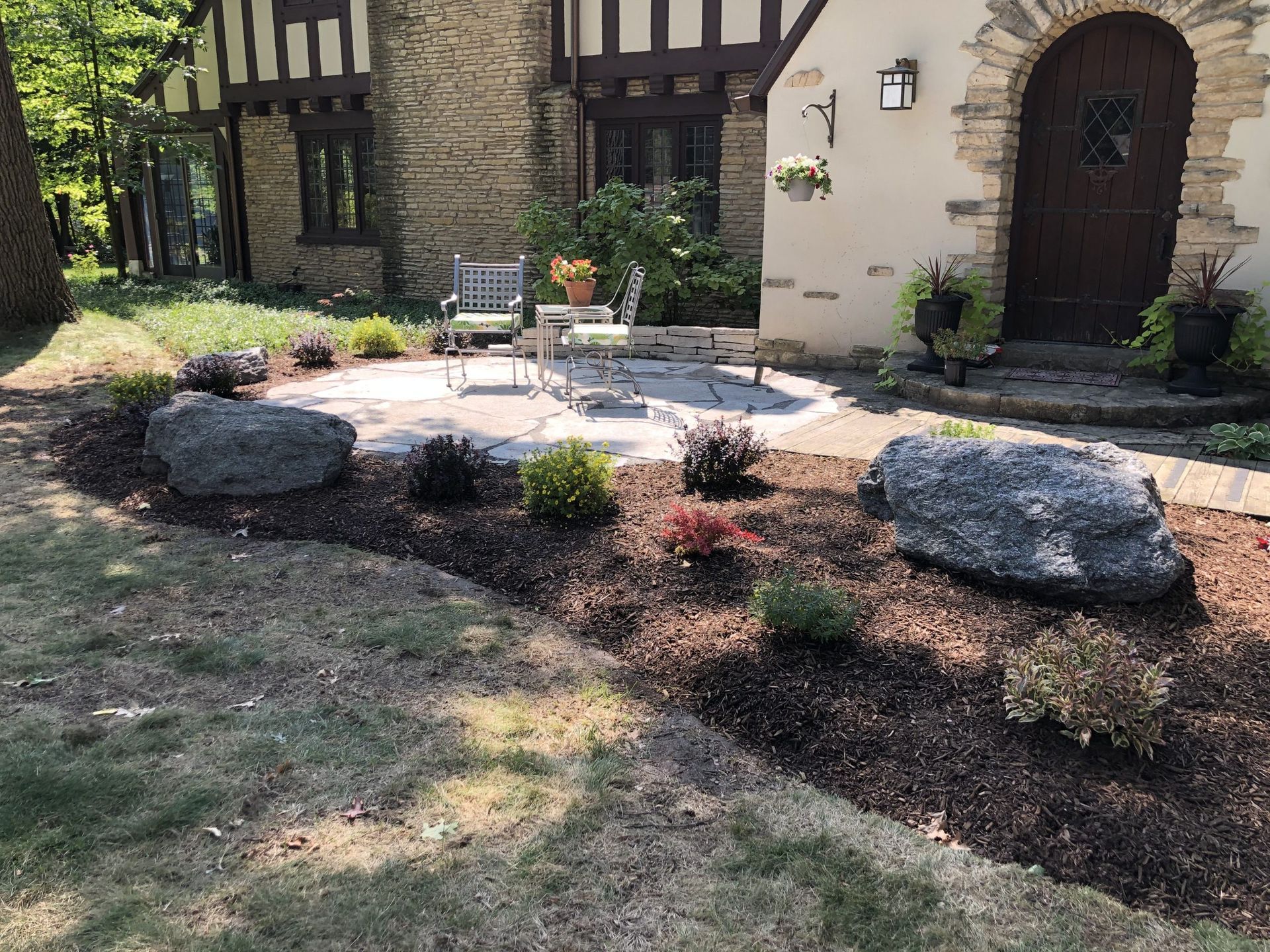 A landscaped front yard with a stone patio, chairs, and large rocks.