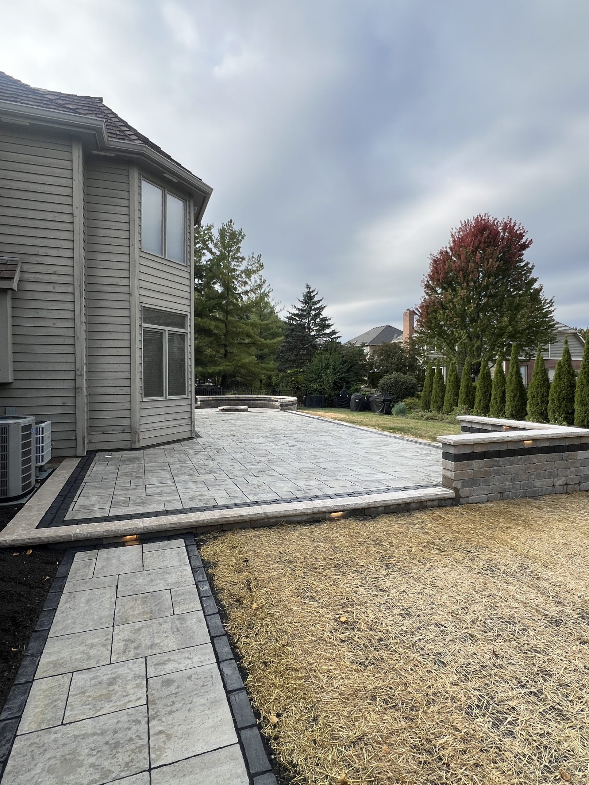 Backyard patio with stone pavers, retaining walls, gravel, and a brick house under a cloudy sky.
