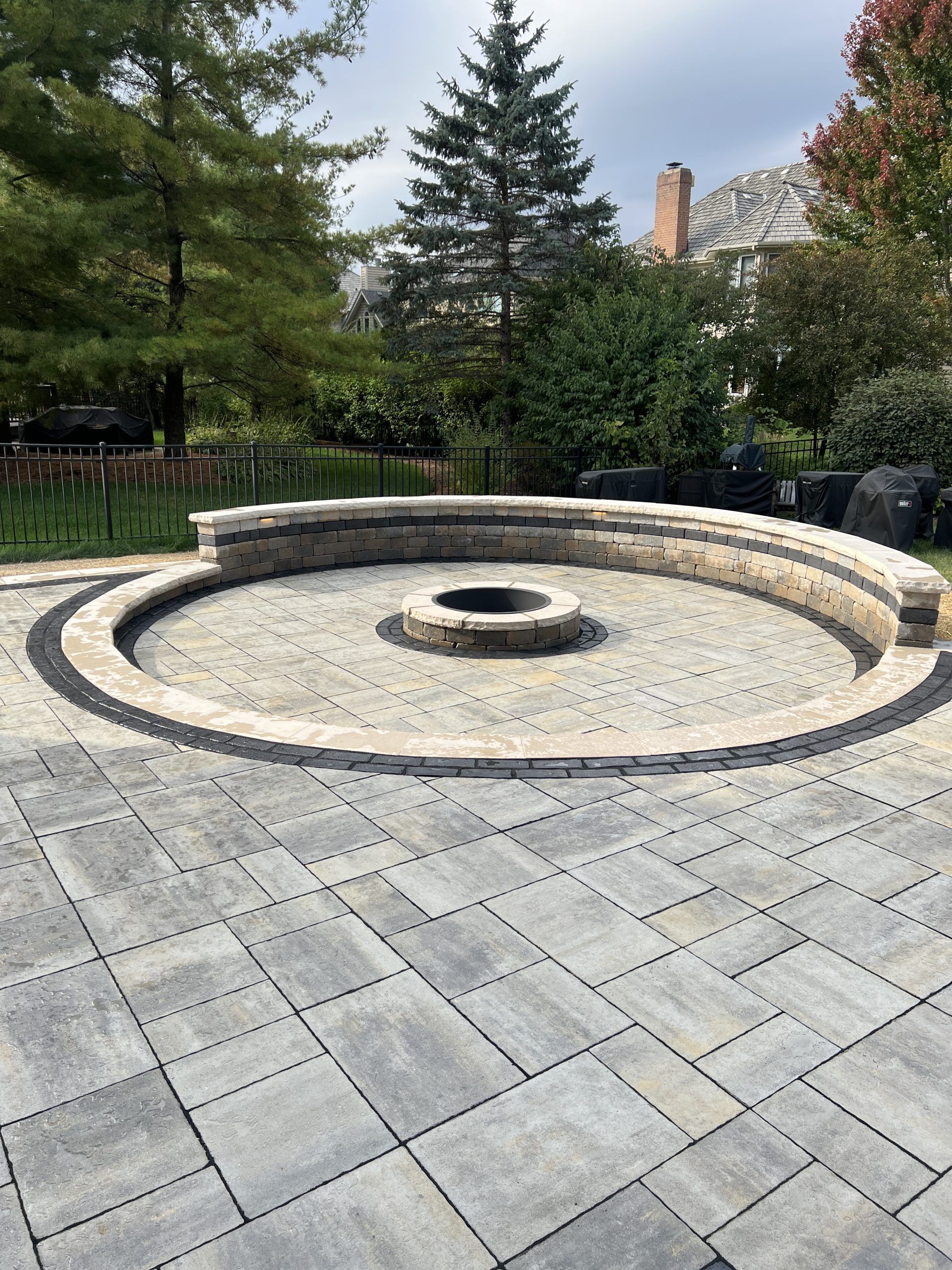 Circular stone fire pit on a paver patio, surrounded by a low stone wall, trees in the background.